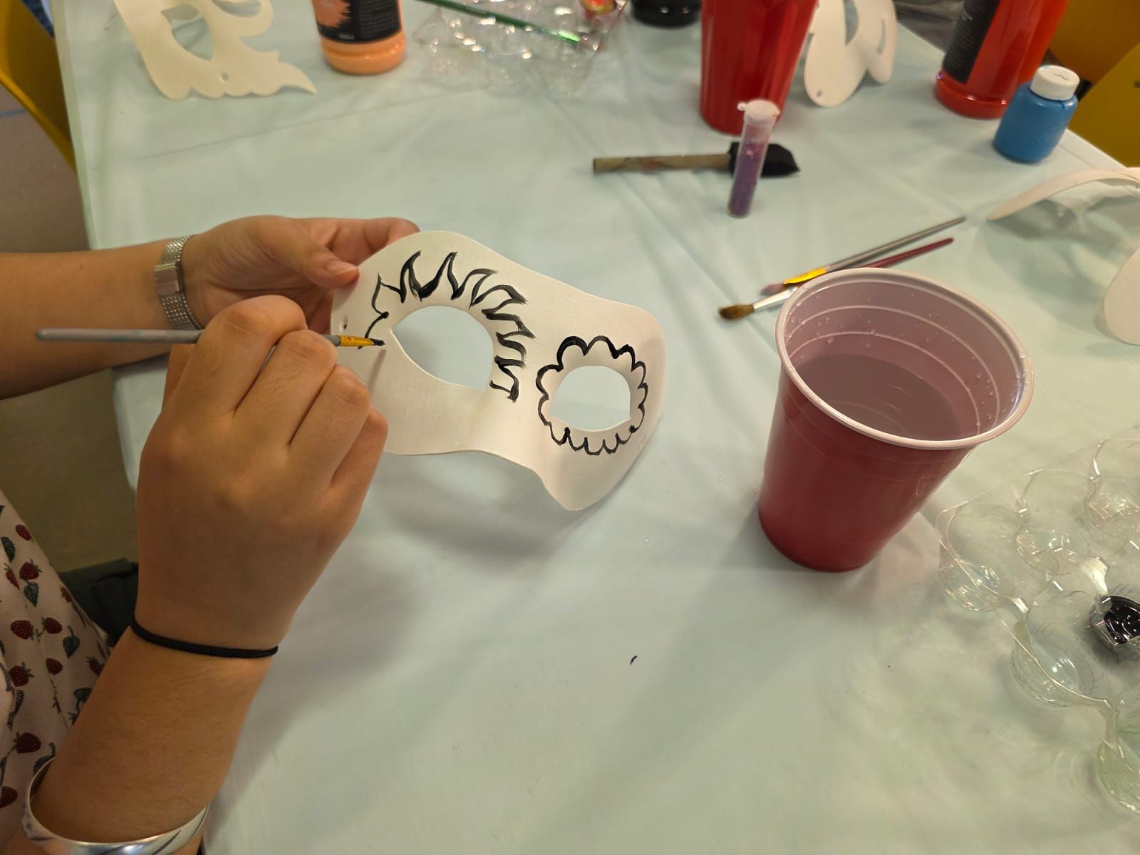 Person painting a white mask with yellow paint at a table. Red cup and paint supplies nearby.