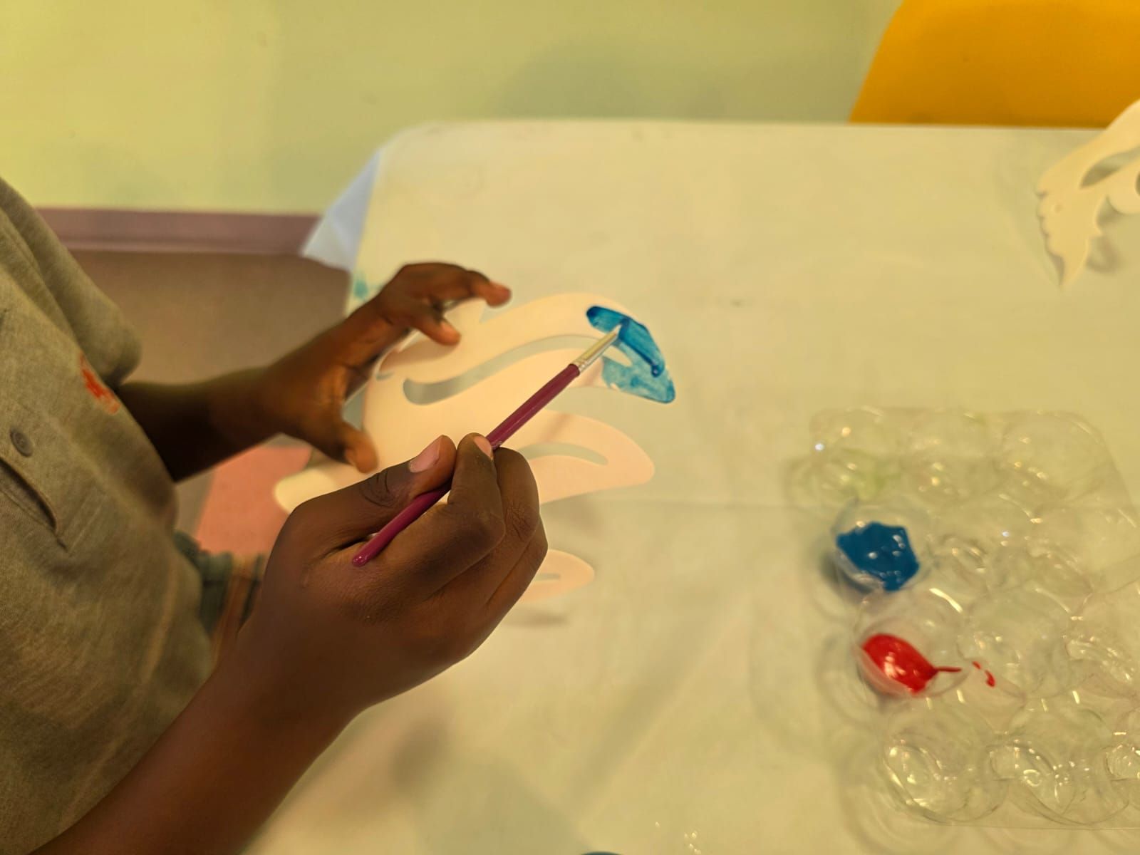 A child paints a white mask with blue paint, using a paintbrush at a table.