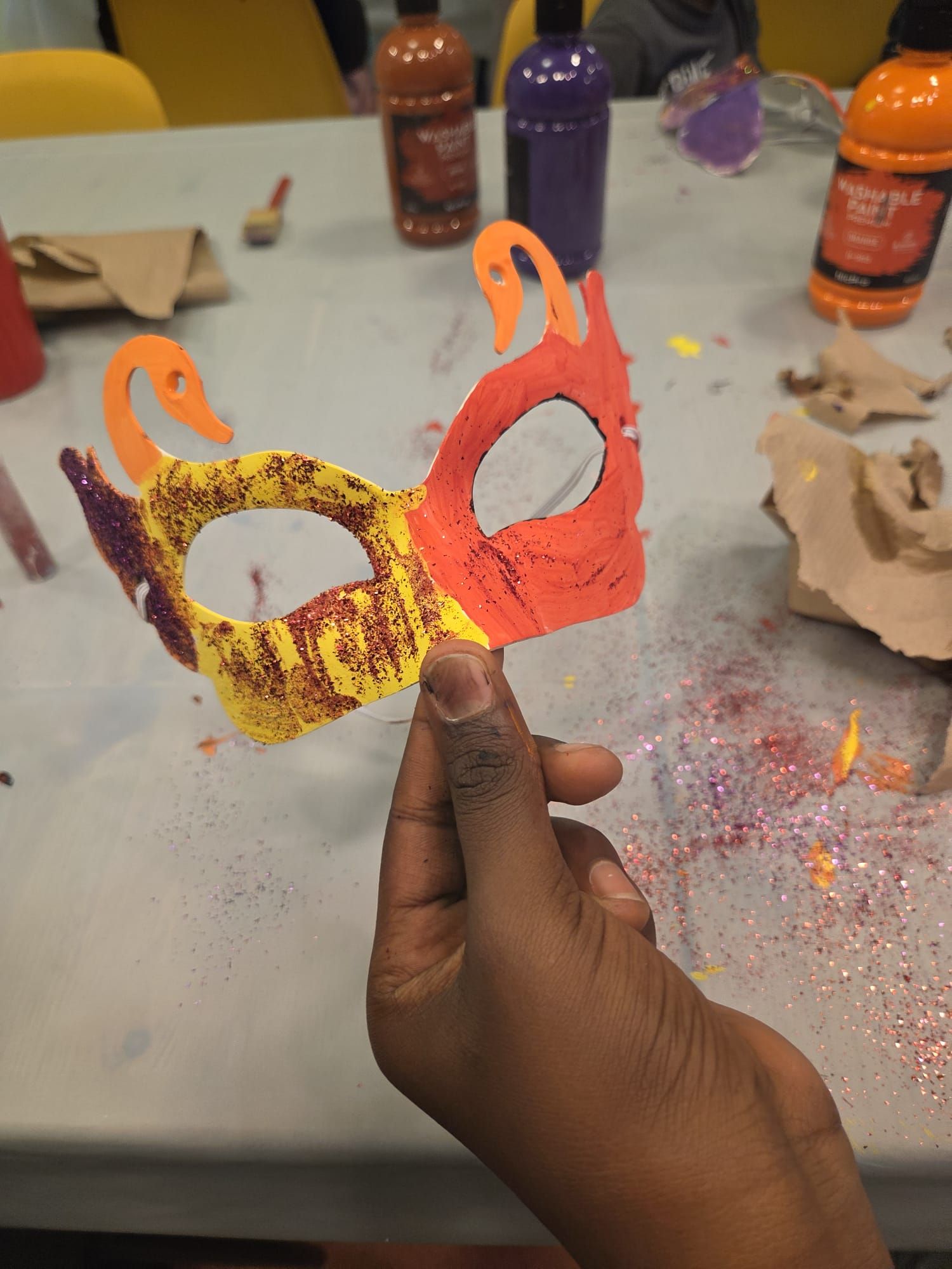 Person holding a colorful handmade mask with red, orange, and yellow paint and glitter on a table with paint bottles.