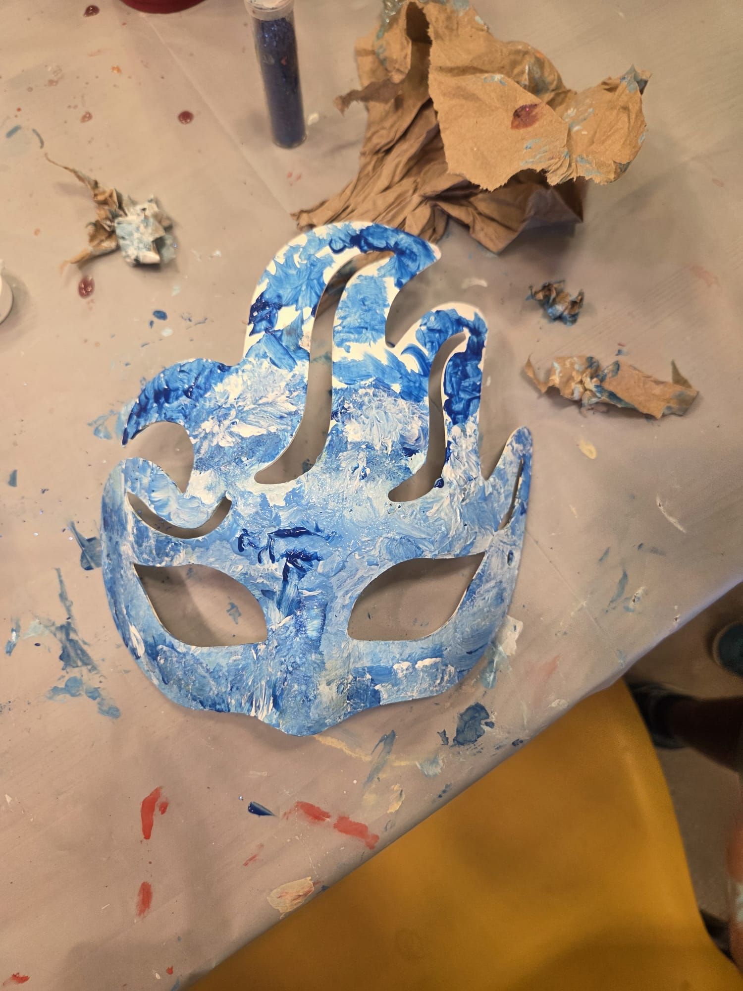 Blue and white painted mask on a table, with other art supplies scattered around.