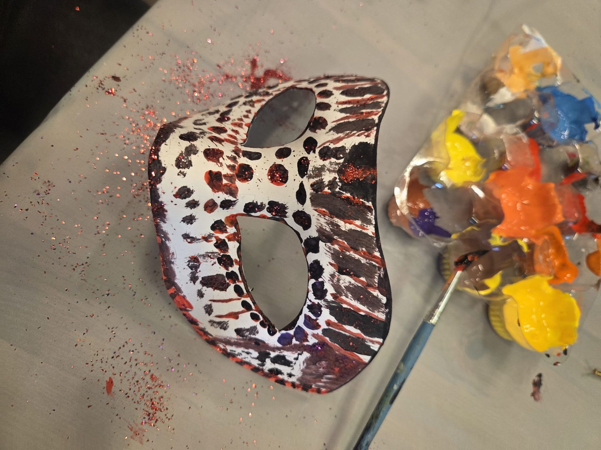 A decorated mask with red and brown patterns, next to a paintbrush and paint palette.
