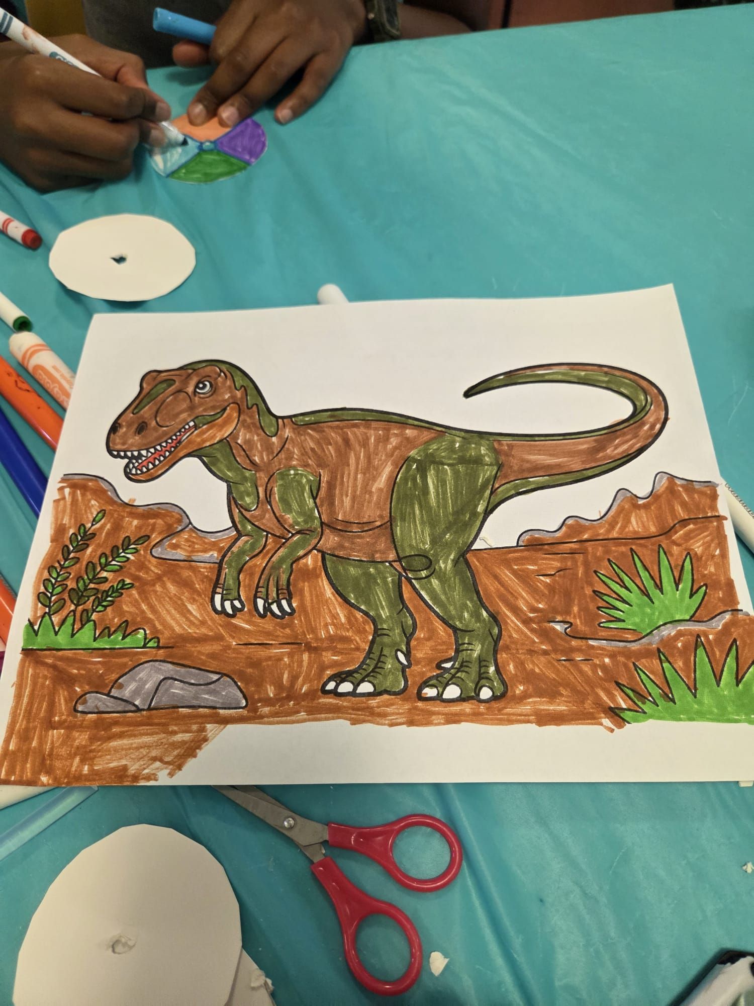 A Tyrannosaurus Rex coloring sheet with brown and green colors, scissors, and someone coloring nearby.