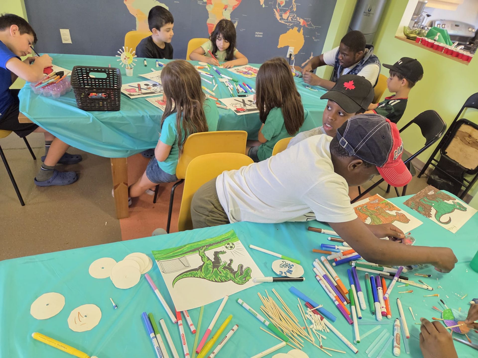 Children drawing and coloring at a table in a classroom; various markers and art supplies are scattered around them.
