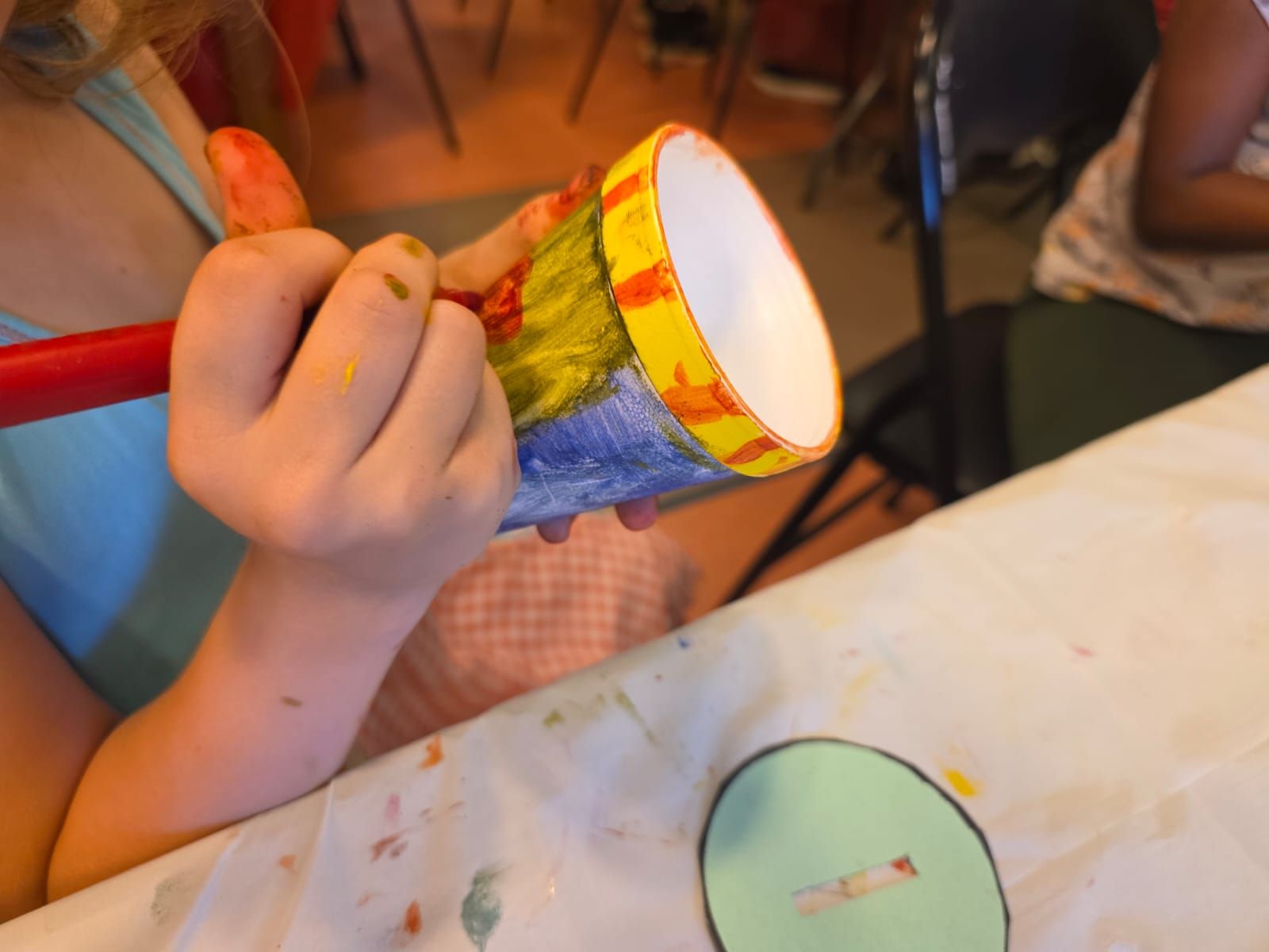 Child's hands painting a drum. Blue, yellow, and red paint. White table.