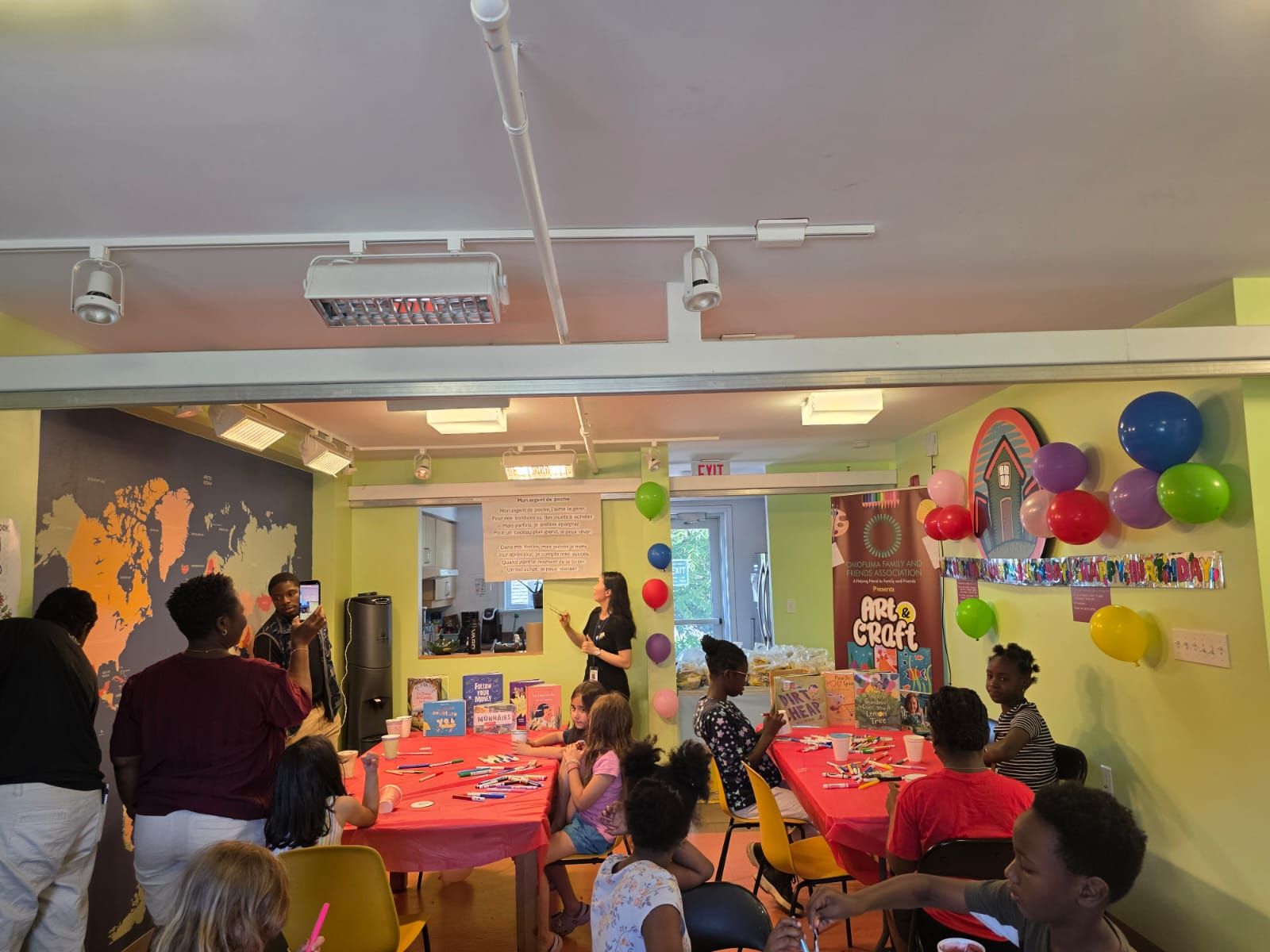 Children and adults in a room with tables, balloons, and a speaker, at a book event.