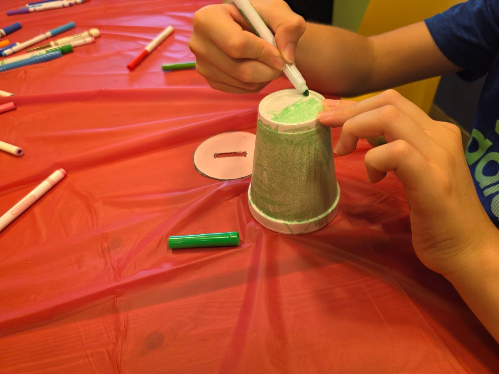 Person coloring a paper cup with green marker at a table, surrounded by other markers.