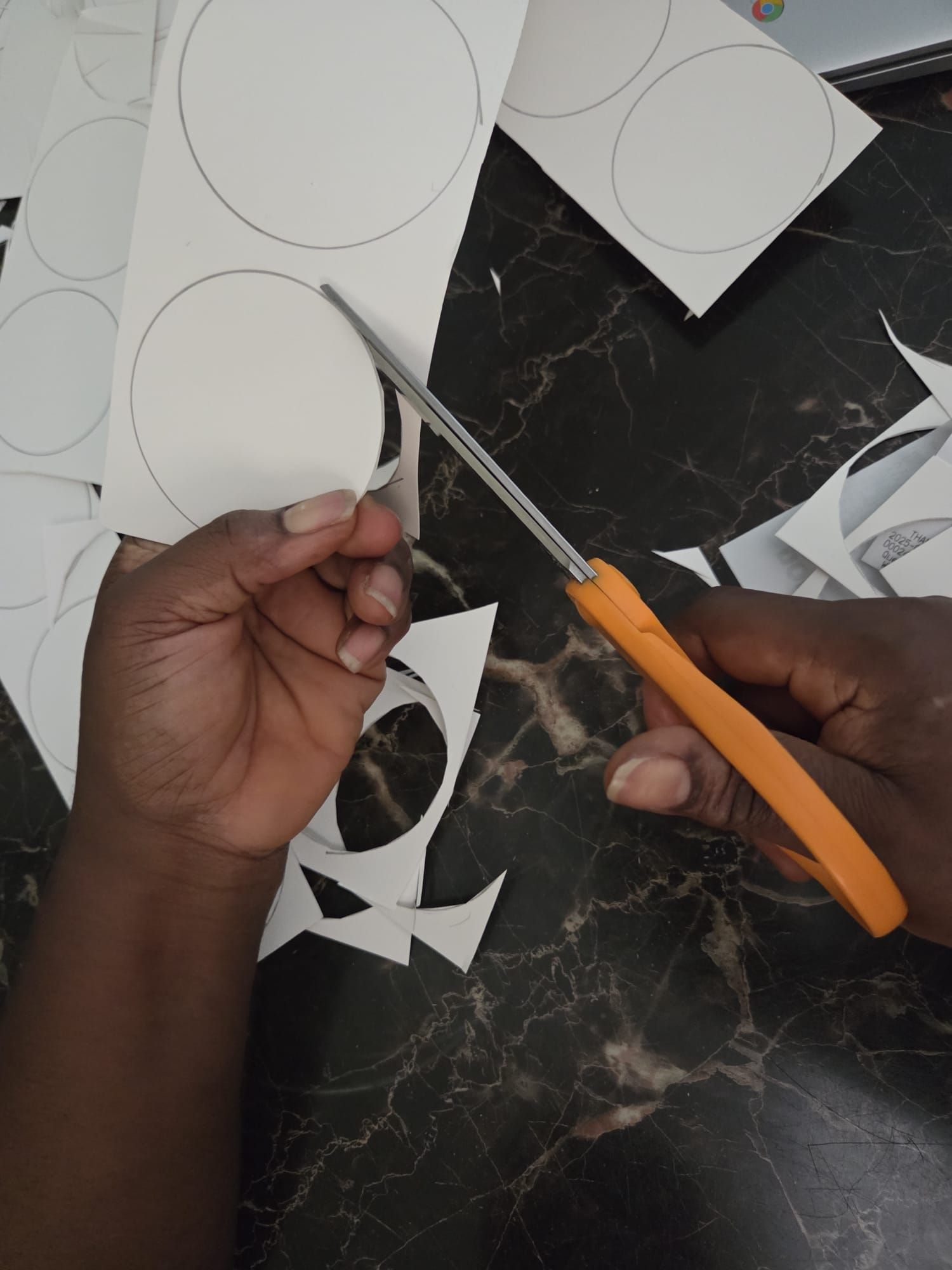 A person with dark skin uses orange-handled scissors to cut out a circle from white paper on a black marble surface.
