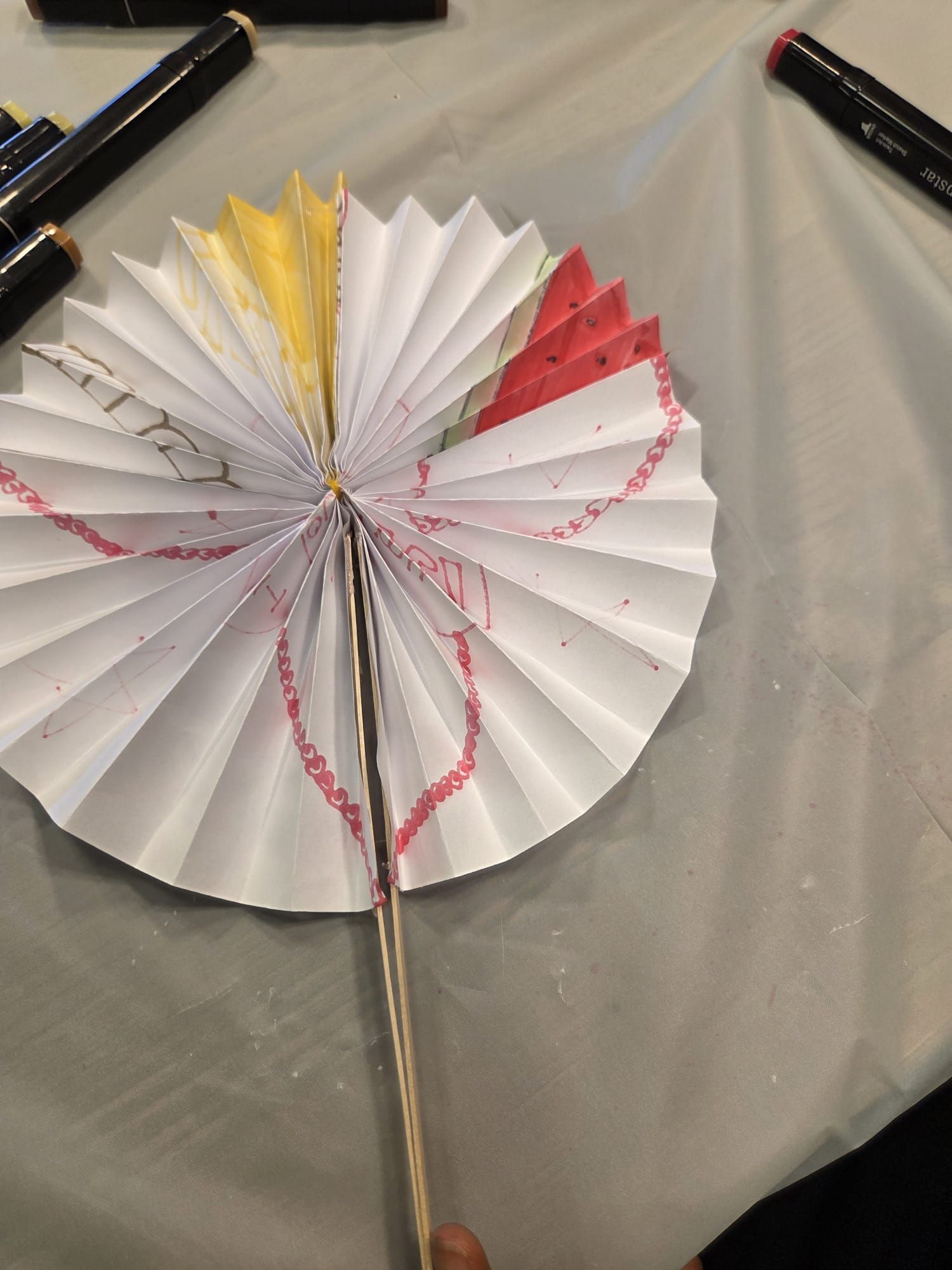 Paper fan decorated with red, yellow, and pink, held by a wooden stick; markers around.