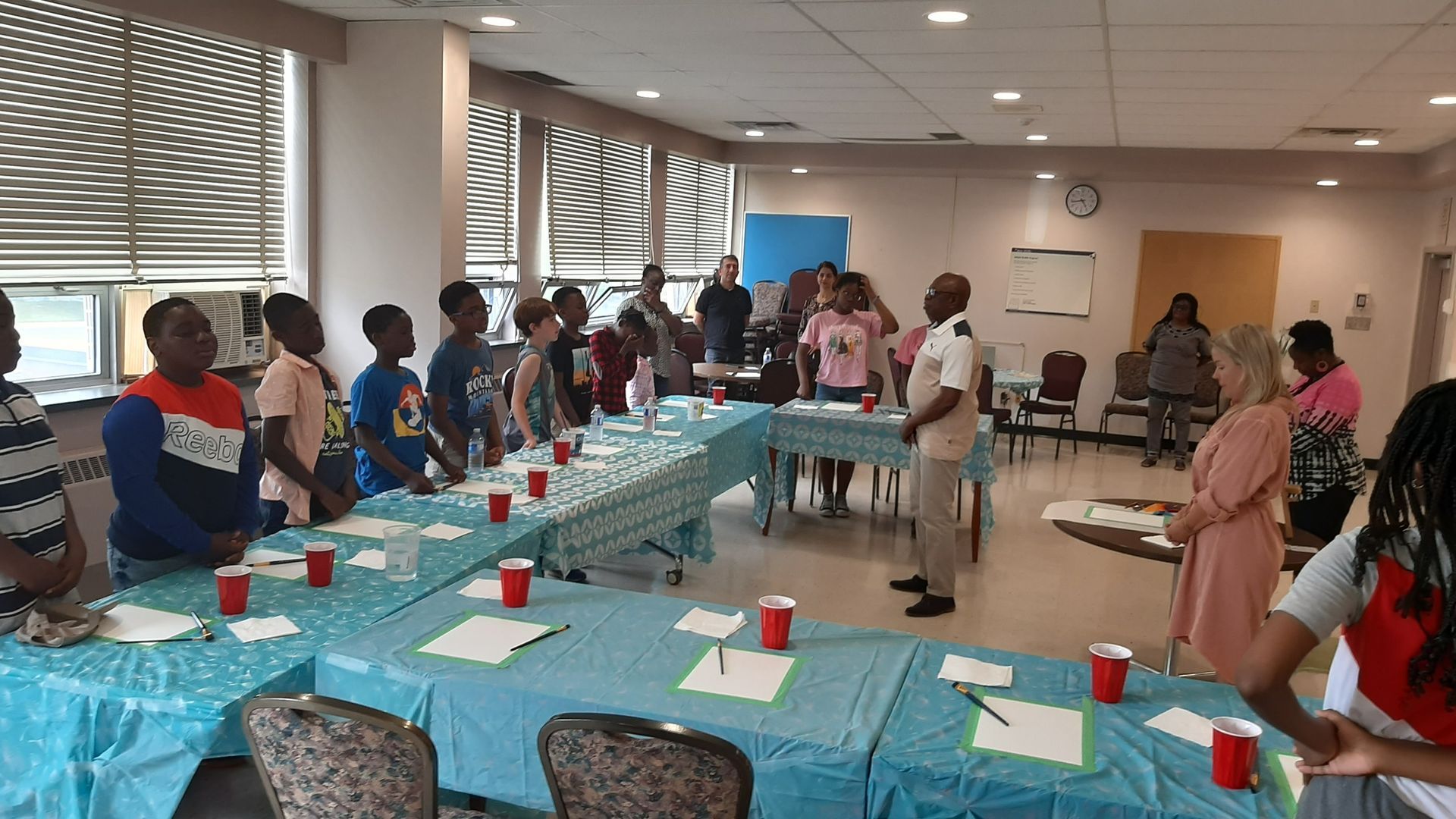 Group stands around tables in a room. People listen to two people standing in front. Red cups and papers on tables.