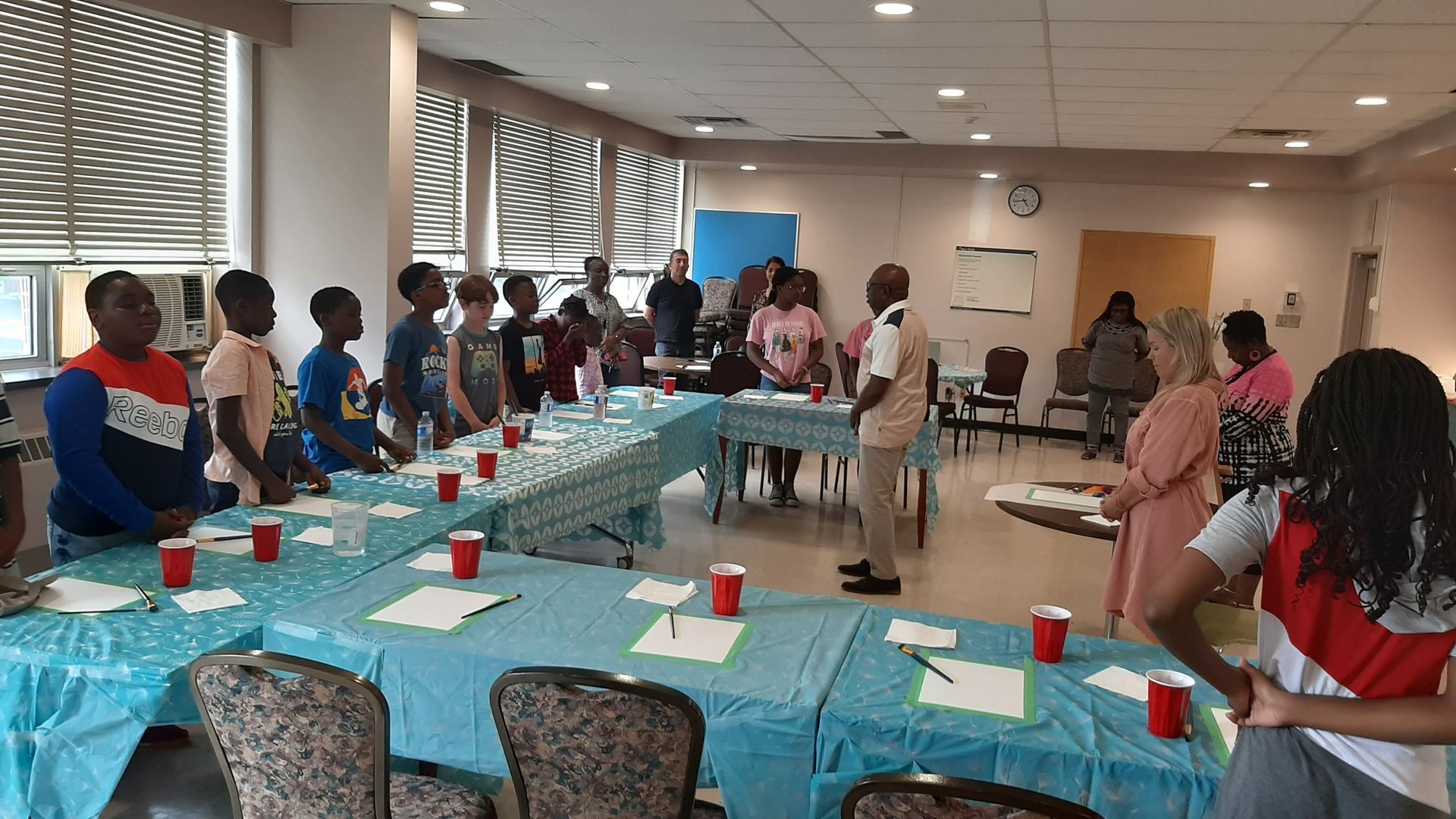Children and adults in a room, standing around tables. Some are holding papers and red cups.