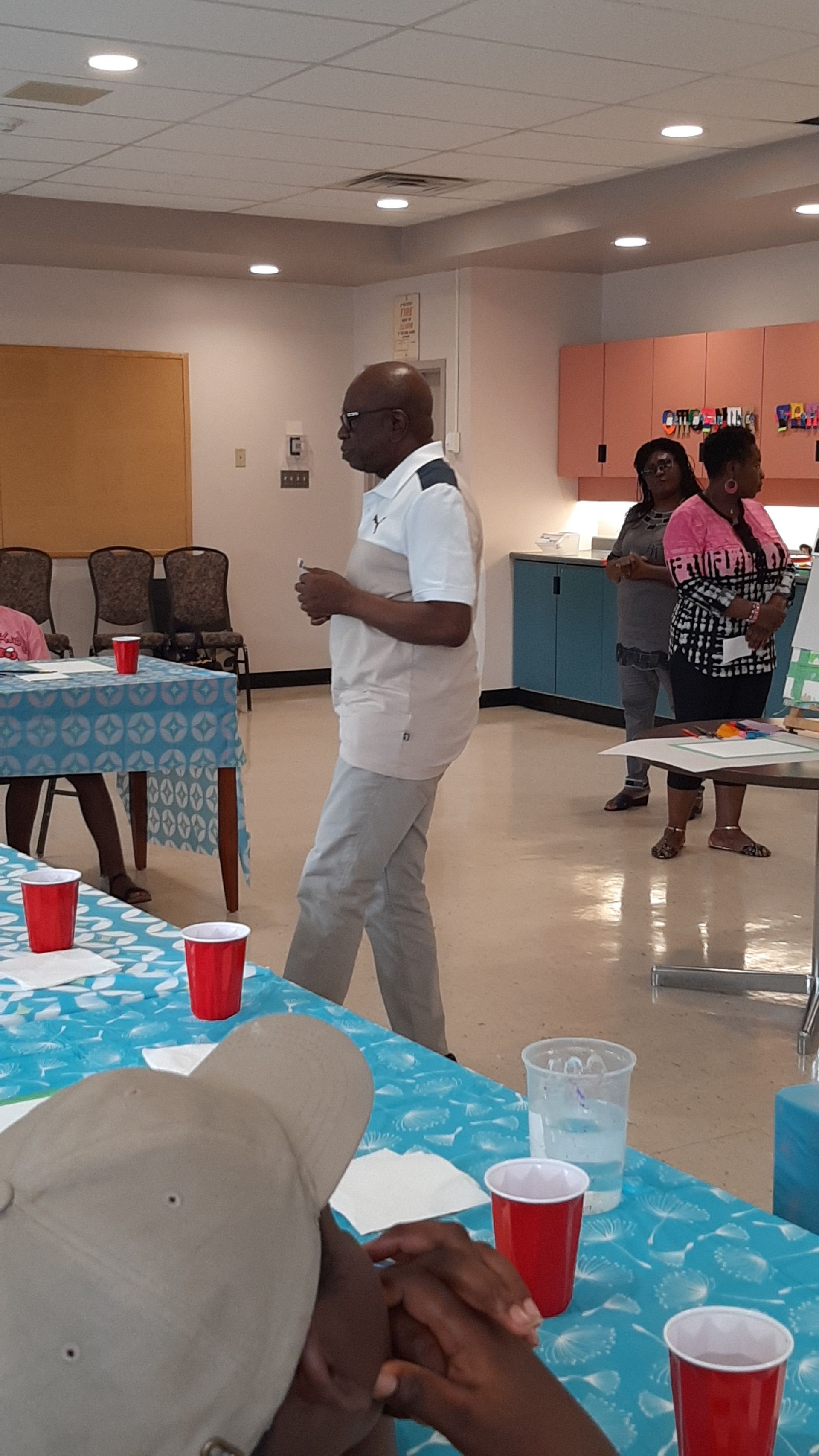 Man speaking, standing in a room with people, tables, and red cups.
