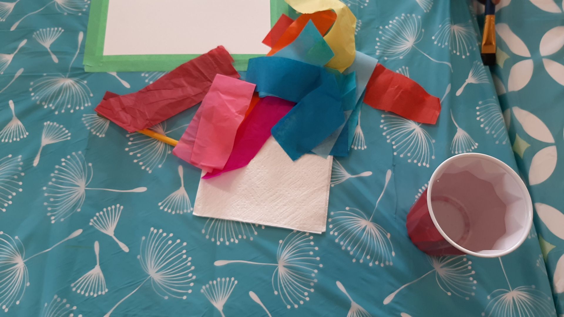 Colorful tissue paper art supplies on blue patterned surface, with cup and paint brush.