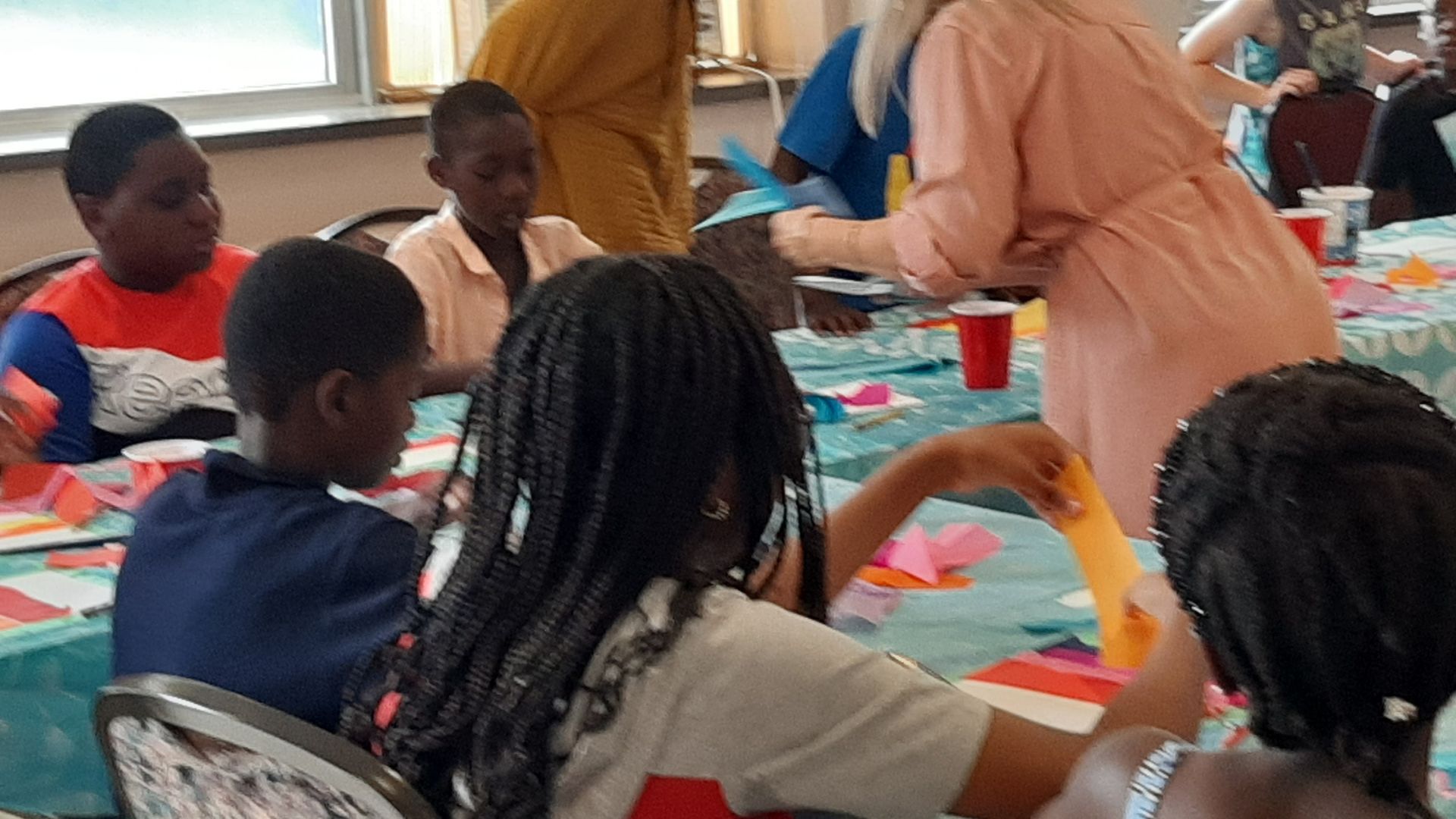 Children and an adult at a craft table, working on projects. Brightly colored paper and supplies.