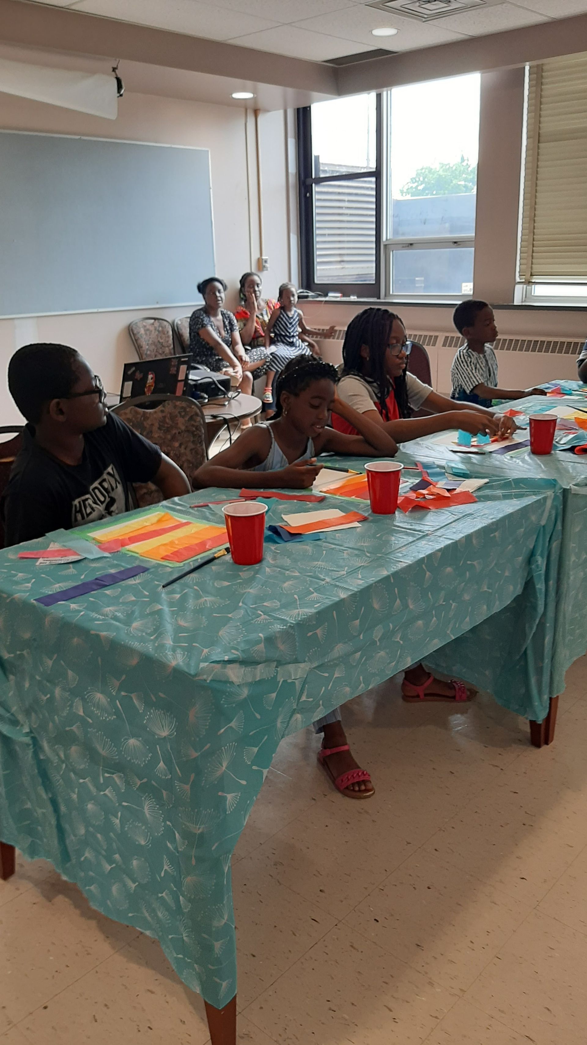 Children at a table with books and red cups, adults in background.