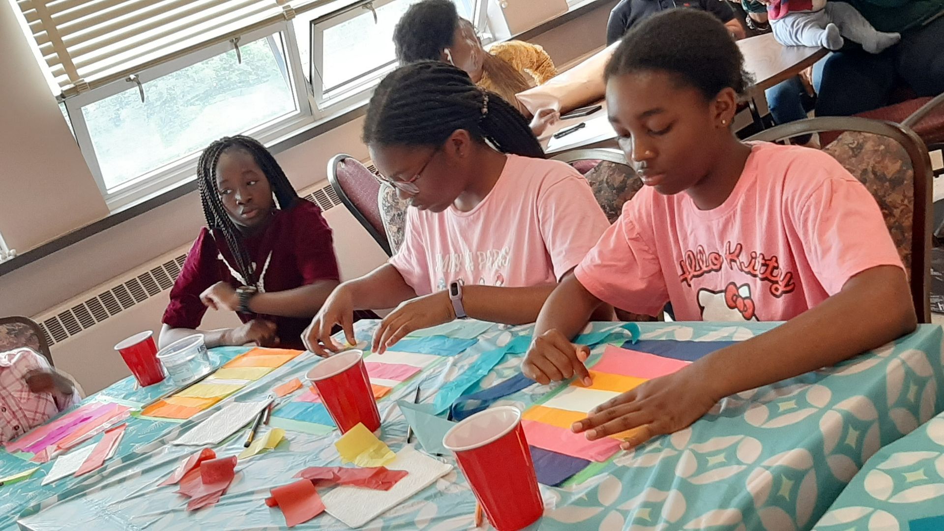 People at a table doing crafts; one person is wearing a pink shirt, red cups are on the table.