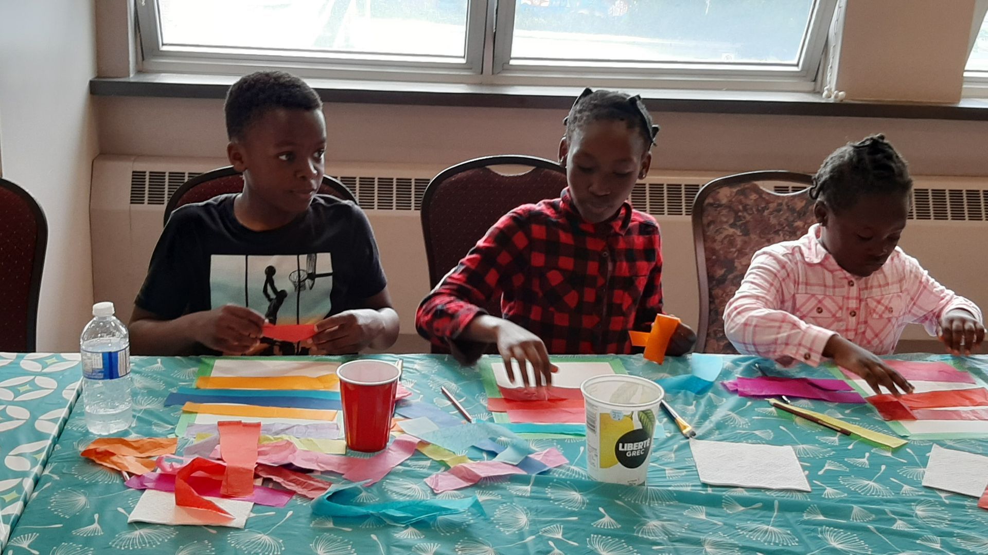 Three children seated at a table, crafting with red and colorful paper.