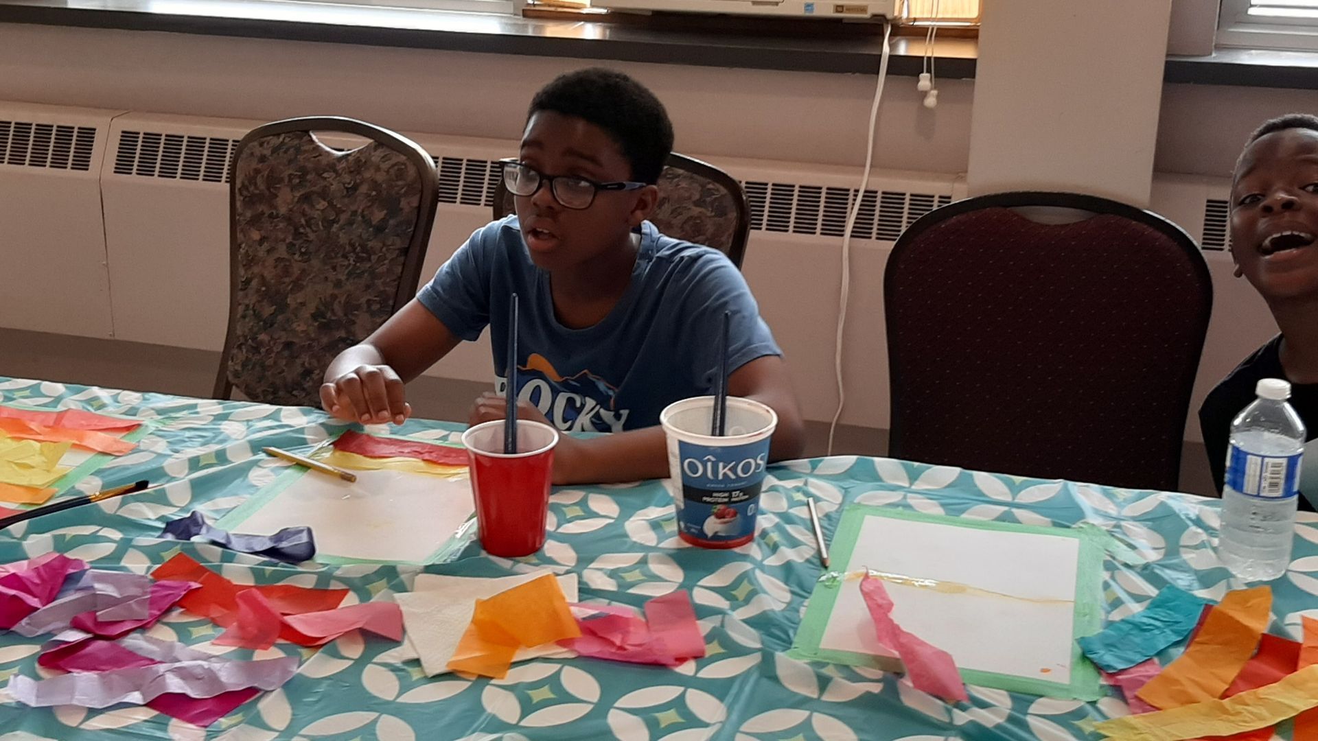 Two boys at a table with art supplies. One boy is focused, holding a pen. Colorful paper scraps and cups are on the table.