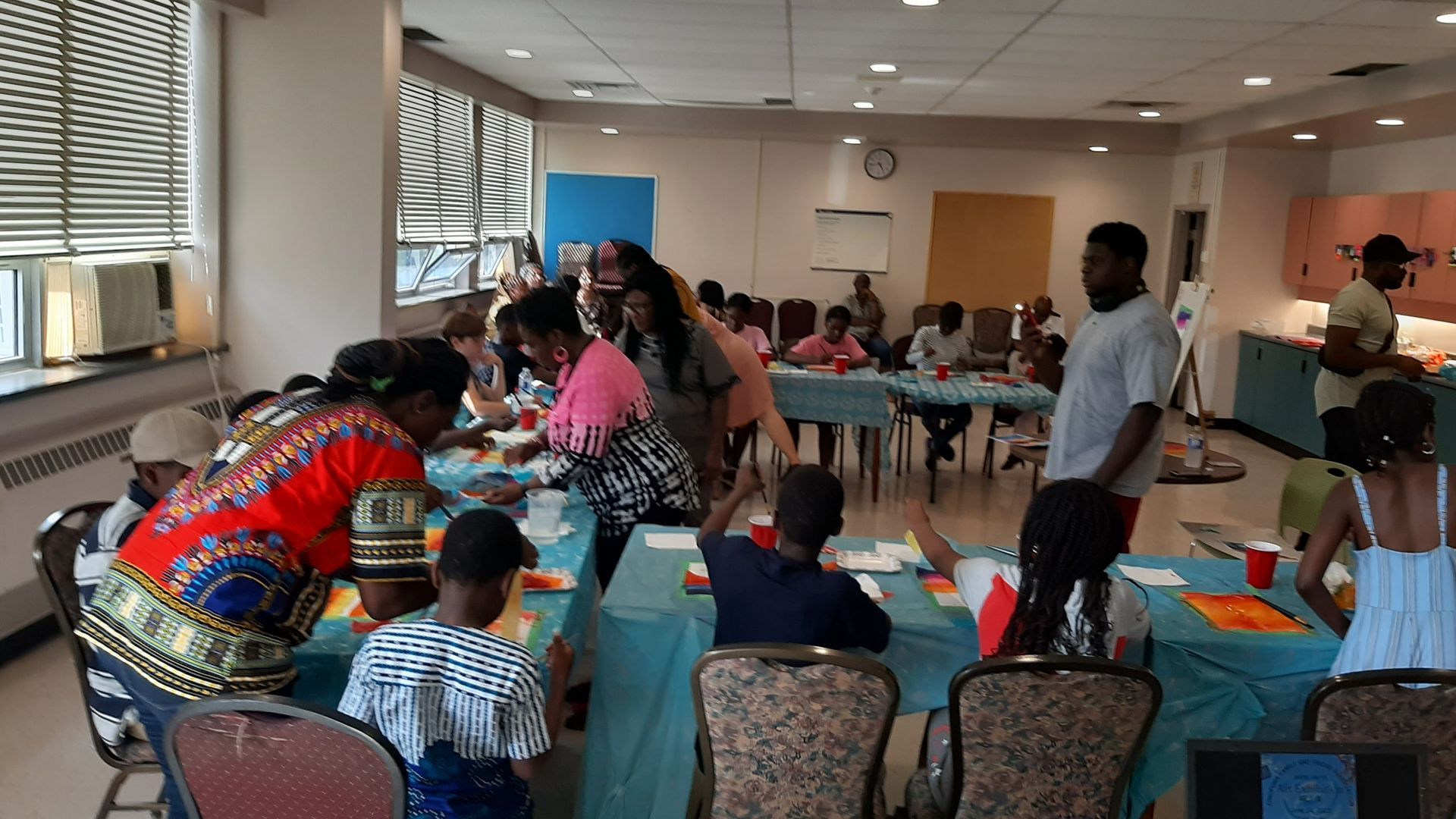 People at tables in a room, some crafting. Several adults and children are present, interacting. Sunlight streams in the windows.