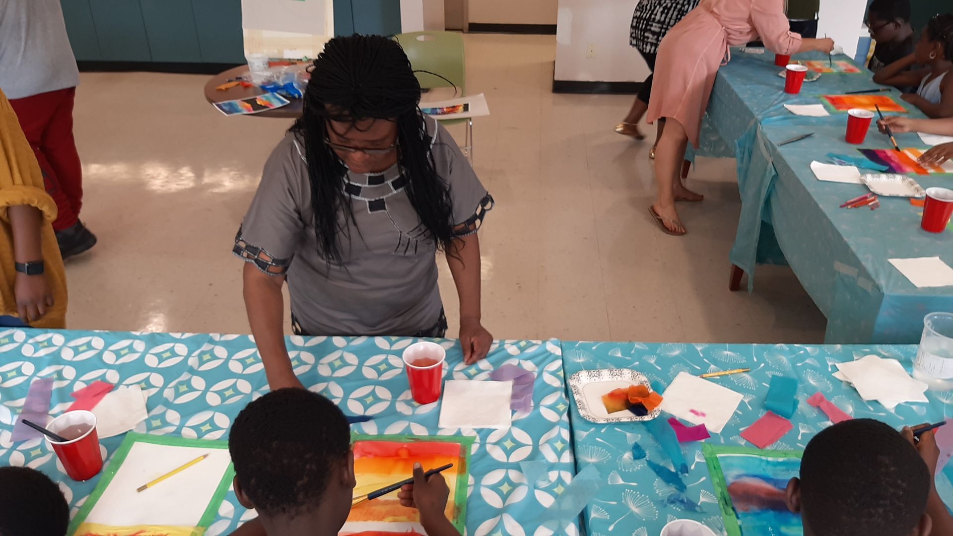 Woman overseeing children at art tables, painting with watercolors, red cups, white paper, brightly colored.