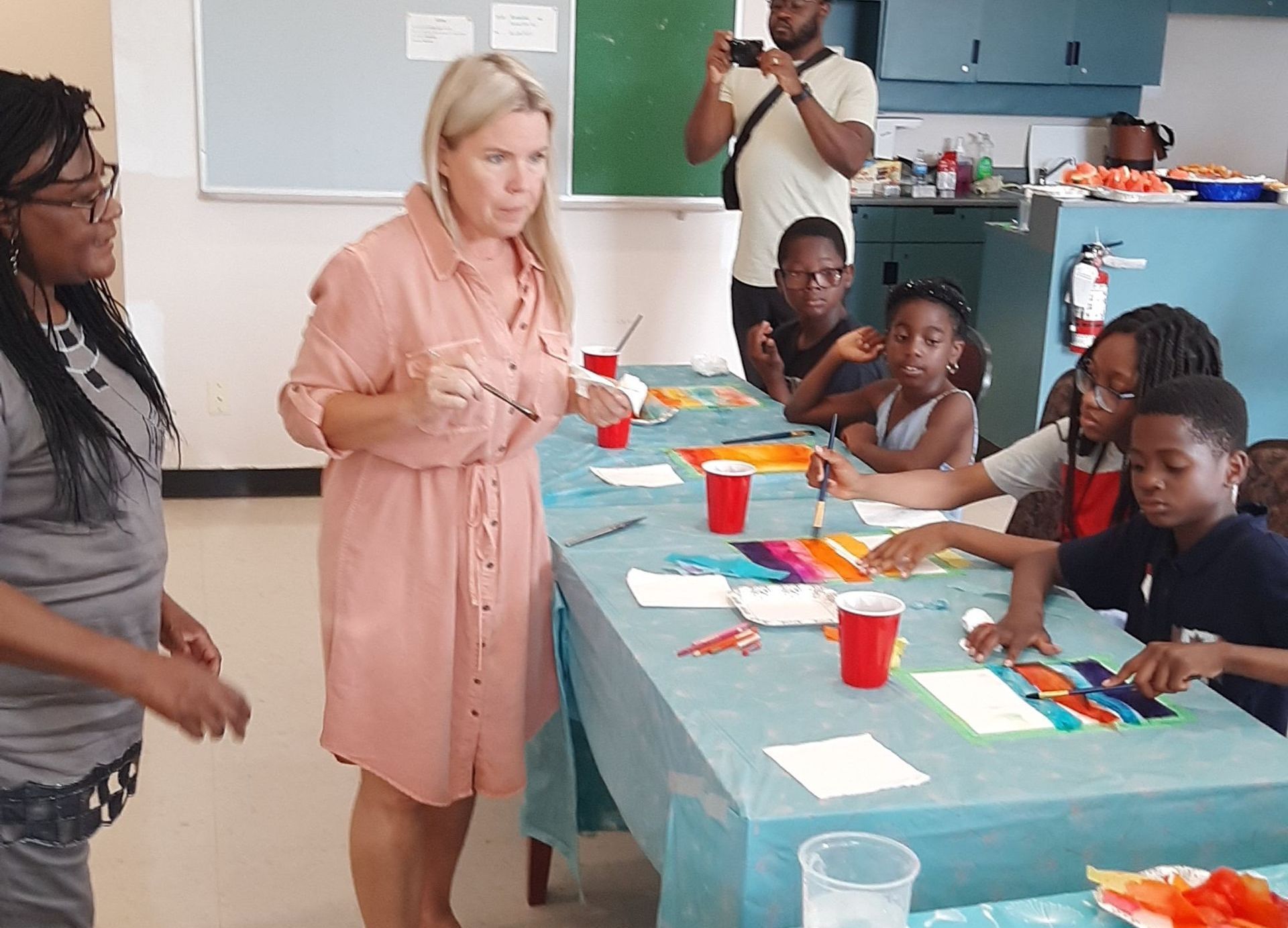 Woman instructs a group of children painting at a table, another woman watches. A man takes photos.