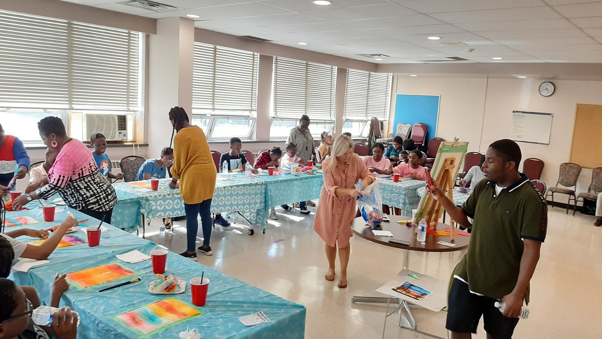 Children at tables painting, guided by adults in a brightly lit room with windows.