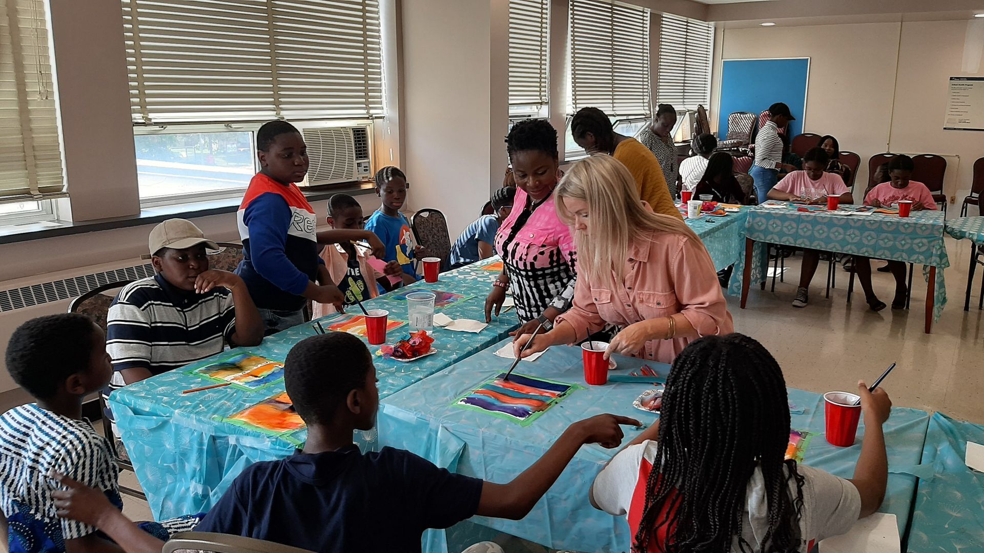 Children and adults painting at a table in a room with a window, painting supplies.