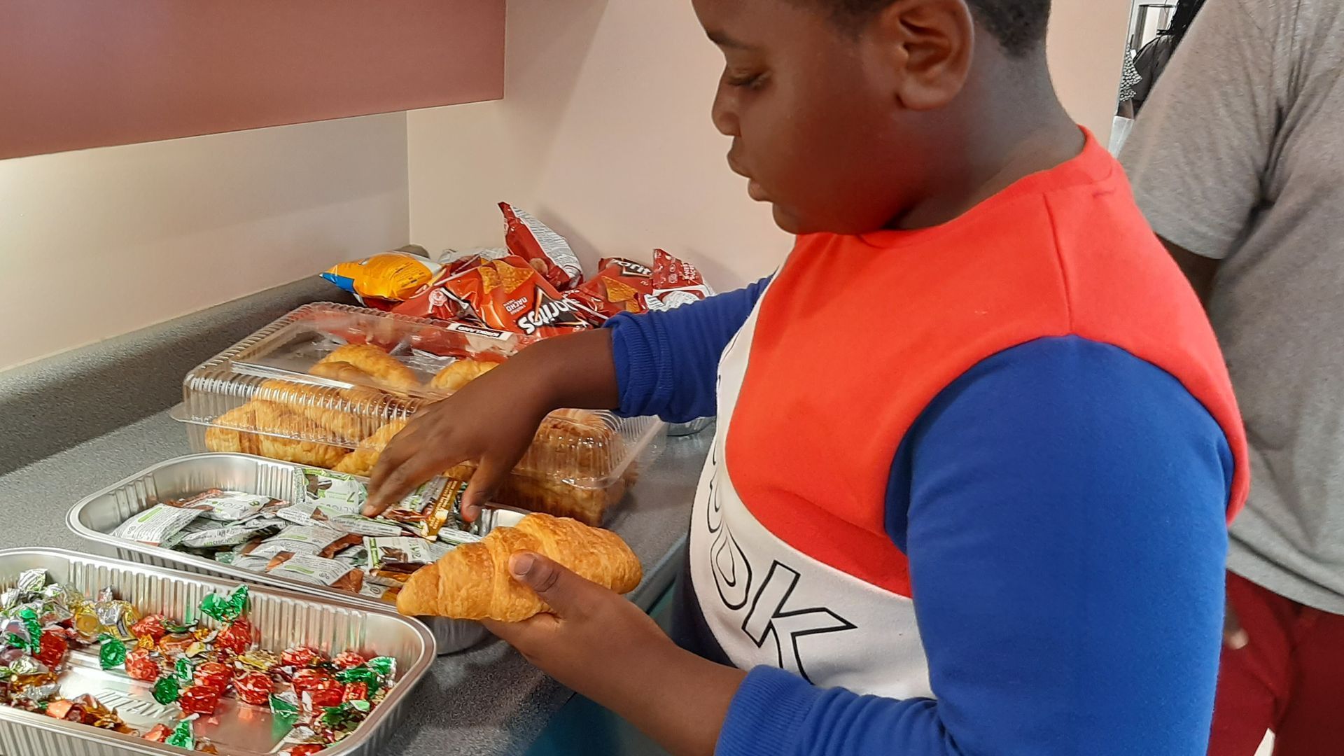 Boy reaching for a croissant at a table with snacks; orange/blue shirt.