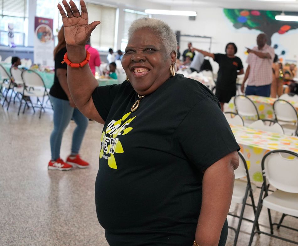 Smiling older Black woman waves from a community event, wearing black shirt, indoors.