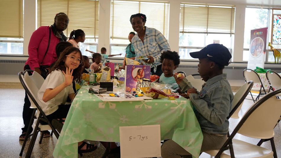 Children at a table, building with toys. Adults oversee. Bright room, sunny, light green tablecloth.