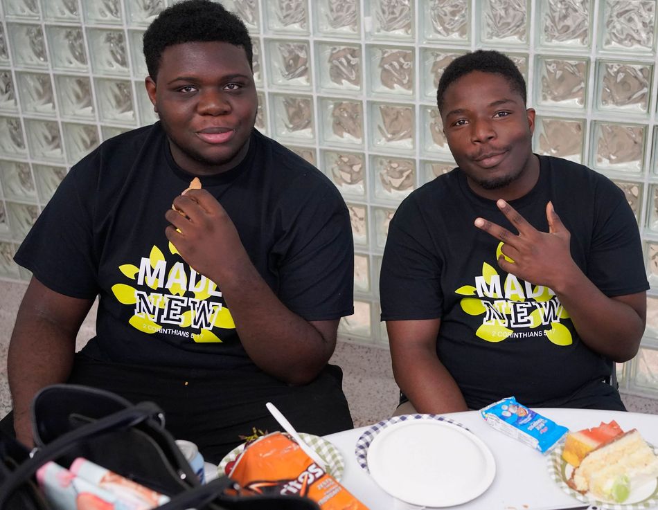Two men in black shirts with logo, eating, at a table with food and a bag.