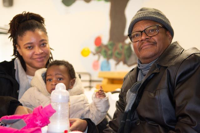 Woman and man with baby indoors. Baby in puffy coat with bottle, man in glasses and hat.