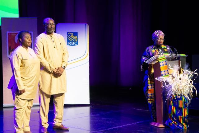Three people on stage: Two in tan outfits stand, one speaks at podium, stage lit with blue/purple.