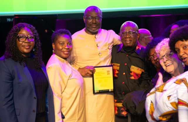 Group of people smiling, holding a certificate. Man in the center, holding award, wearing tan.