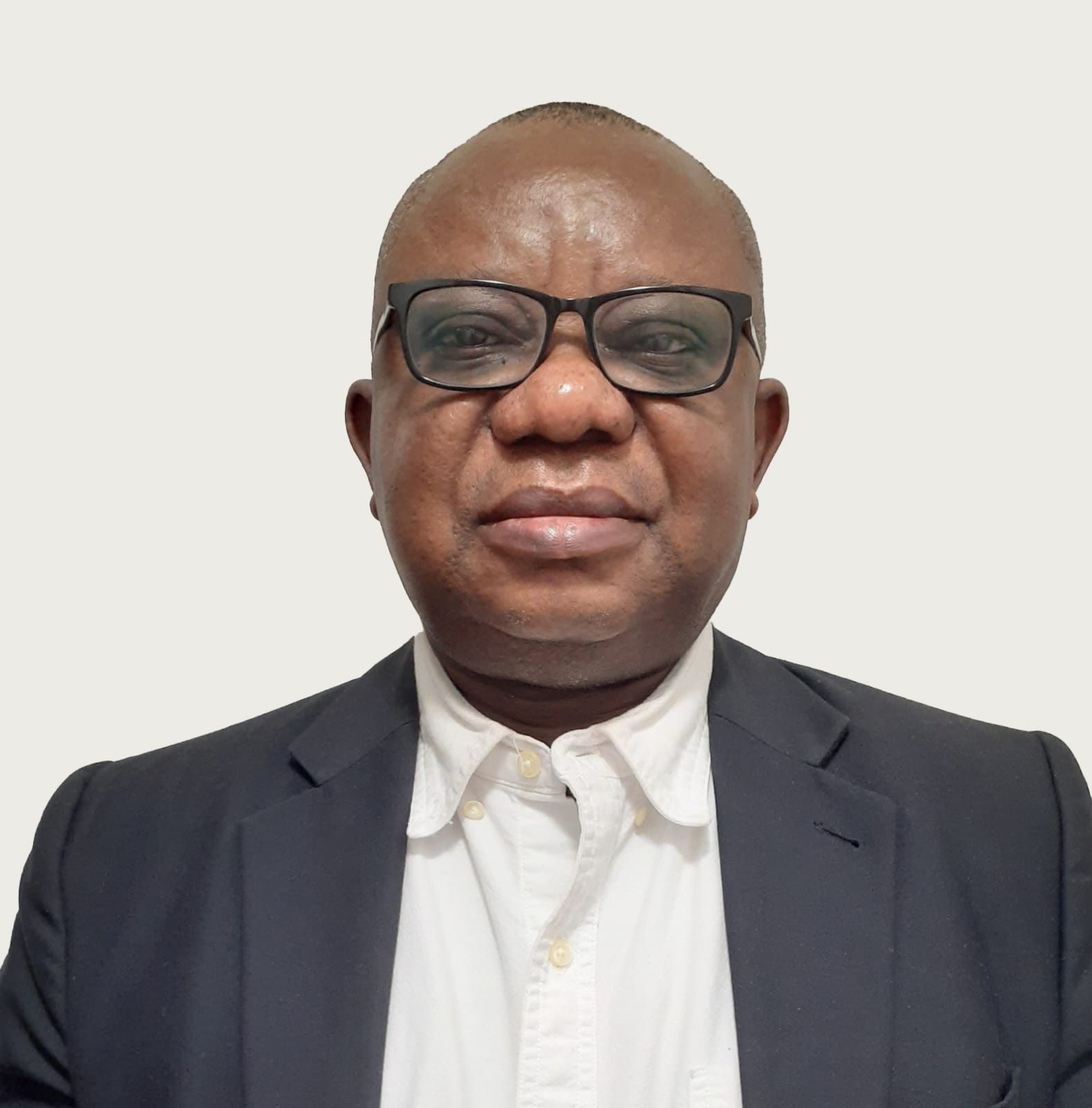 Man wearing glasses, white shirt, and dark blazer looking at the camera. Neutral background.