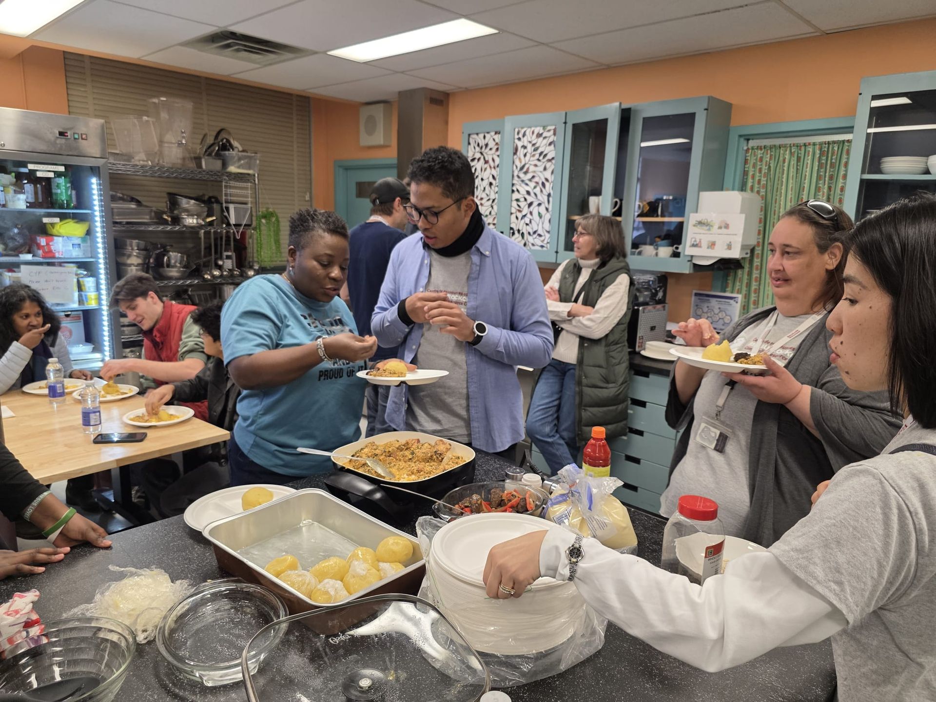 People in a kitchen tasting food from plates; some gathered around a table and counter.