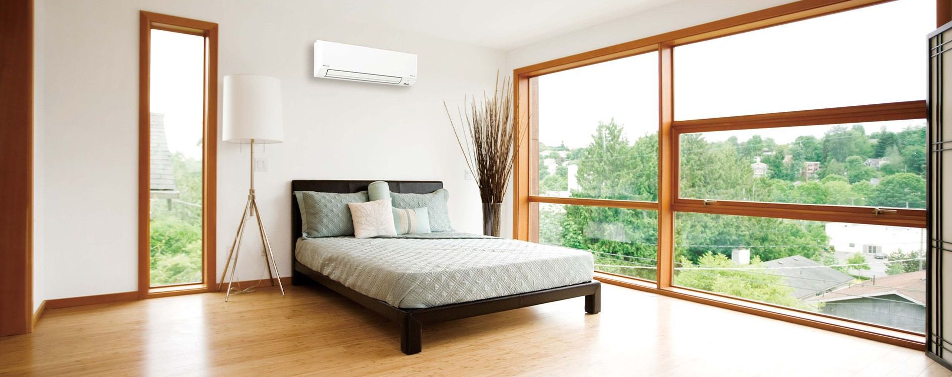 Daikin Alira heat pump installed over a bed in a bedroom with windows overlooking surrounding neighbourhood/trees.