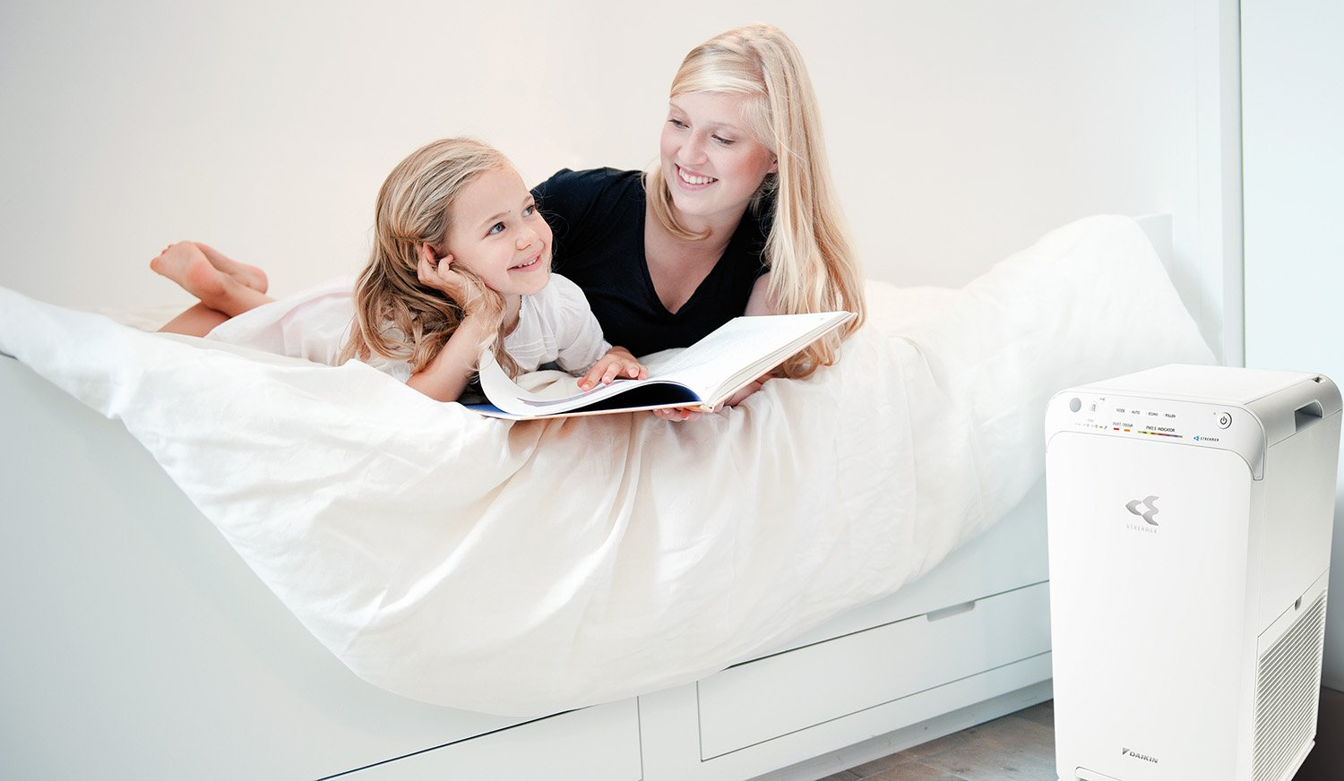 Mother and daughter looking at a book lying on a bed with a Daikin air purifier in the foreground.