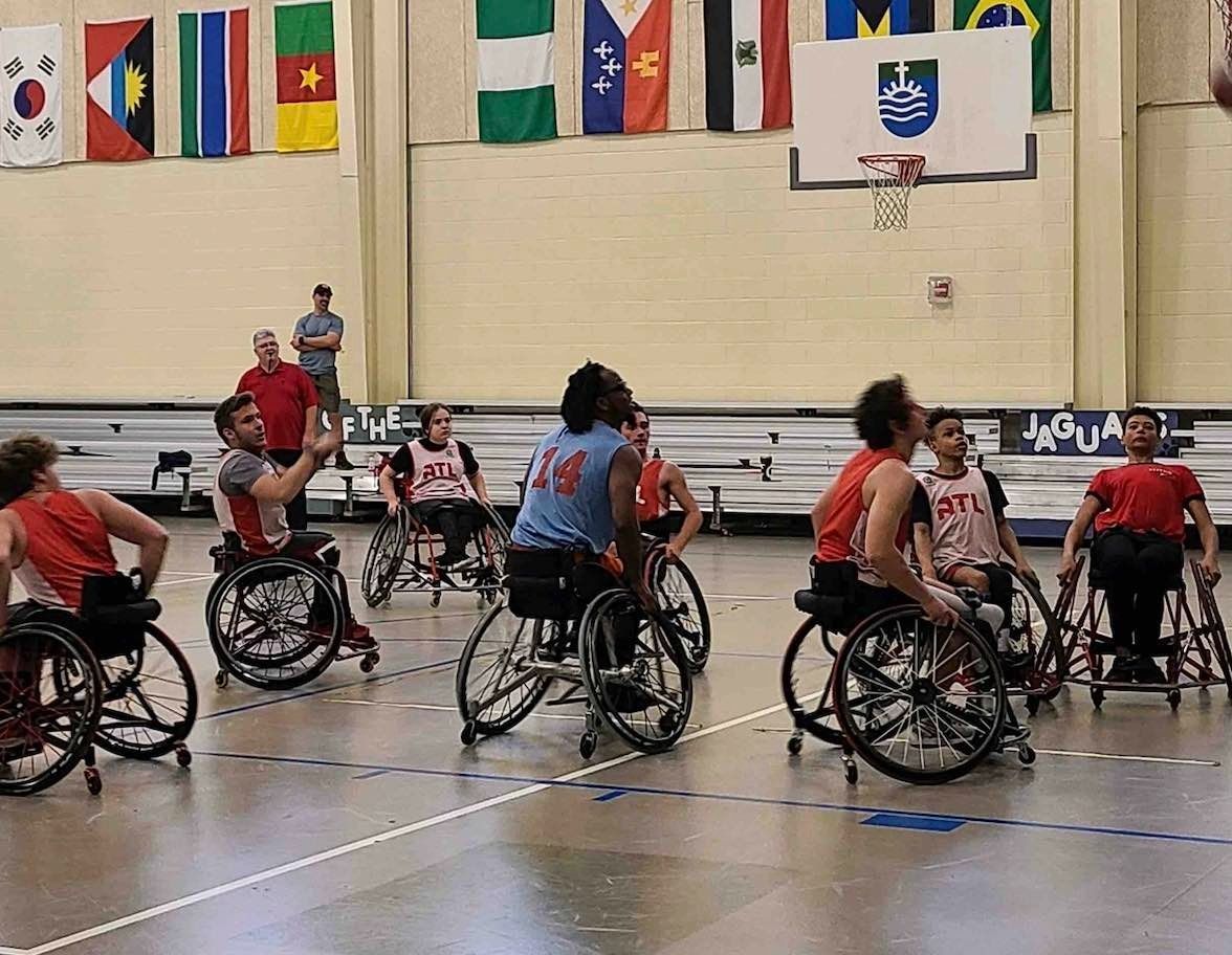 Indoor wheelchair basketball game in a gym, with players in red and blue jerseys under hanging flags.