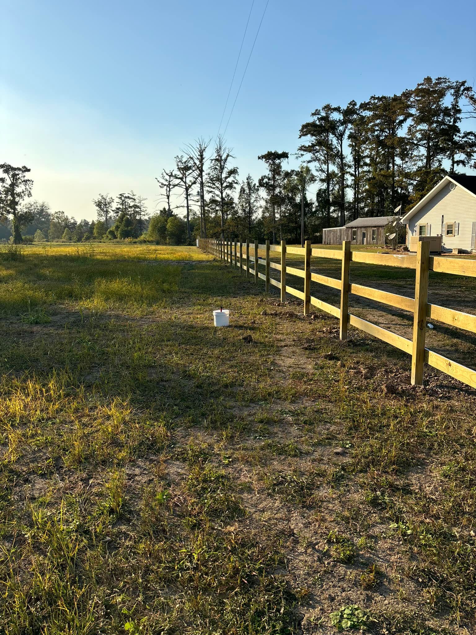 Sunlit grassy field beside a wooden fence and small house under a clear blue sky