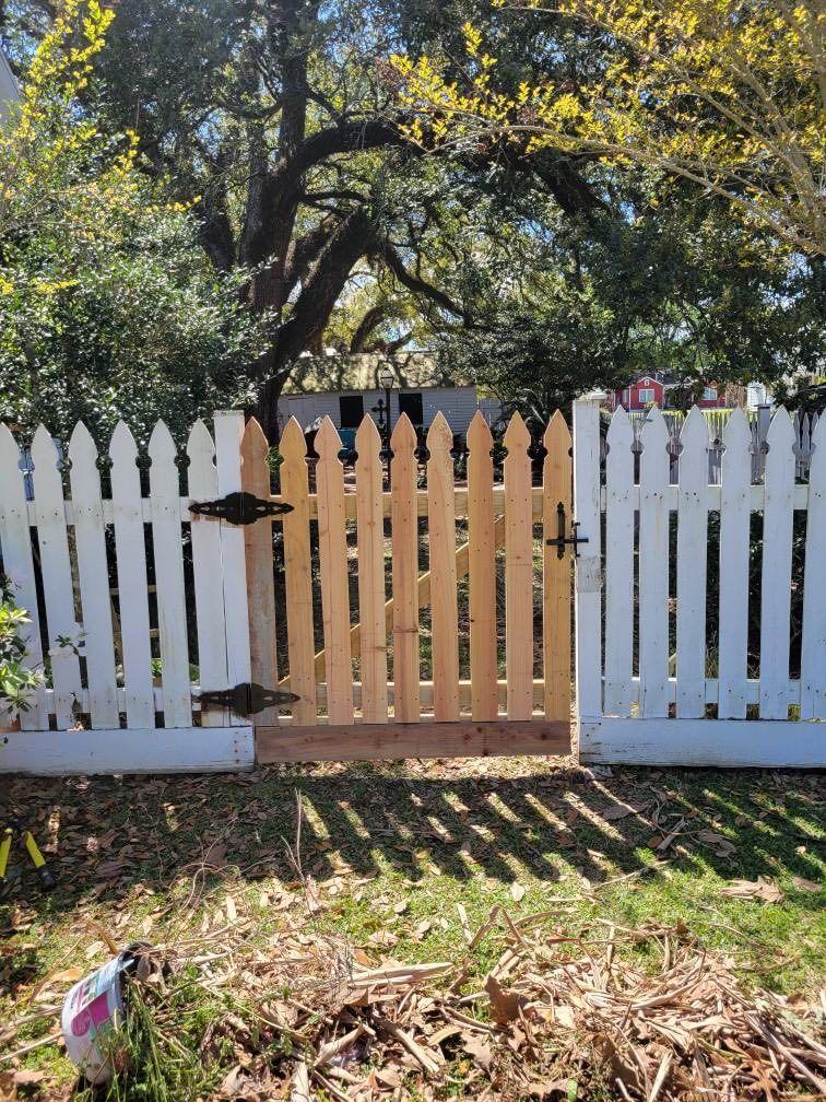 White picket fence with a tan wooden gate in a sunny yard beneath a large tree.
