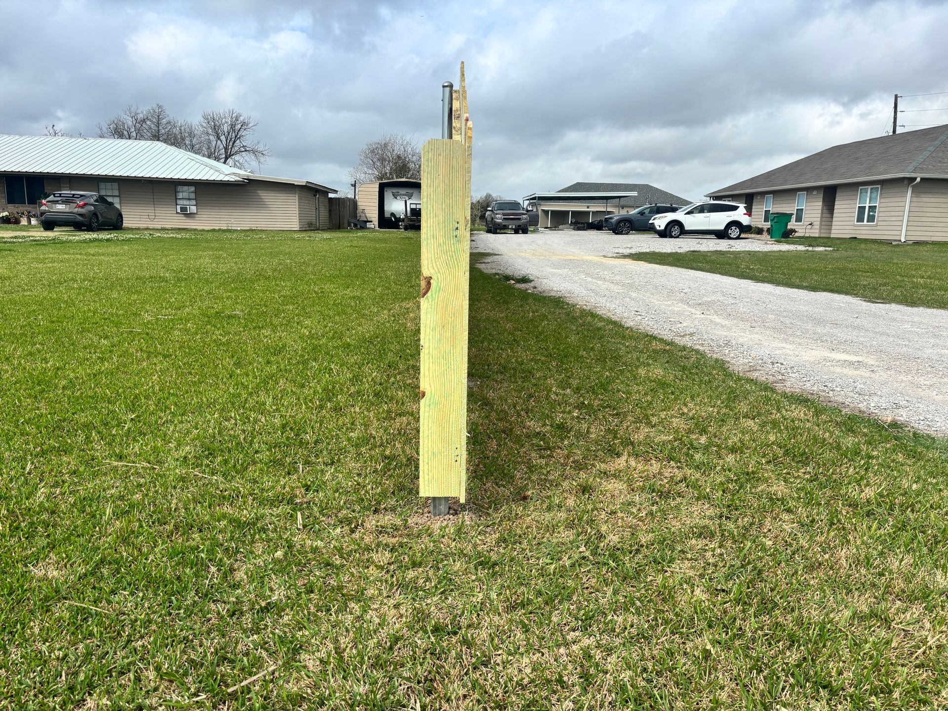 Wooden utility post in a grassy yard beside a gravel road, with houses under a cloudy sky.