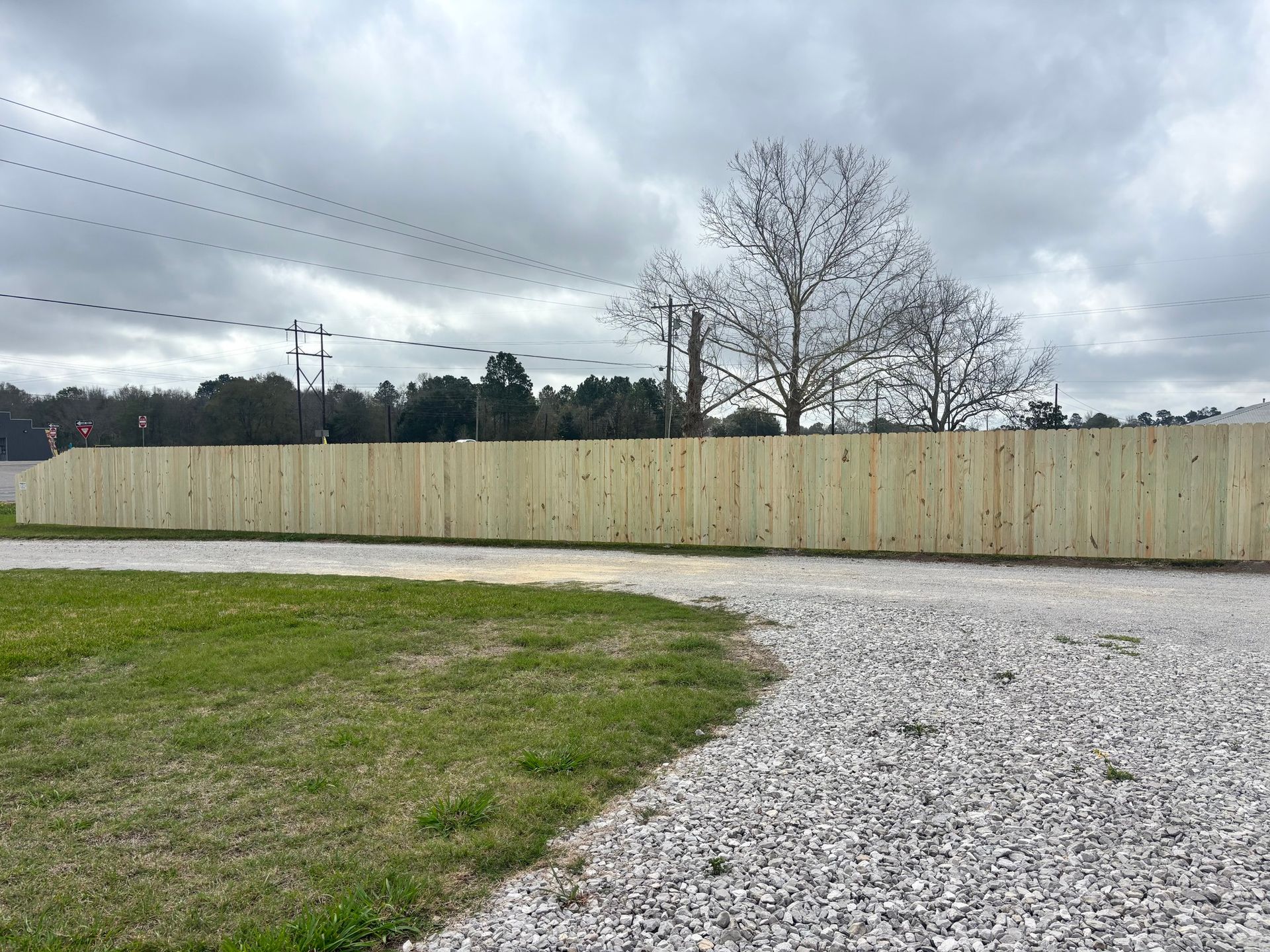 Gravel path beside a beige fence under a cloudy sky, with grass and trees in the background