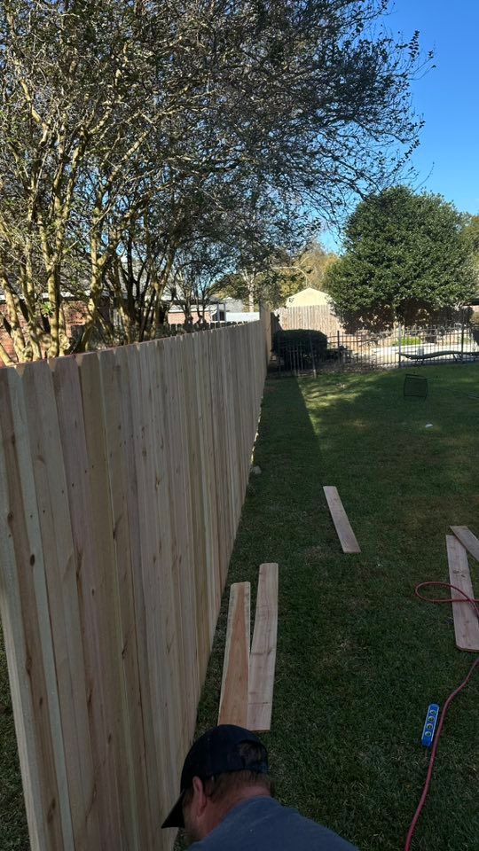 Man working along a wooden fence in a grassy backyard, with trees and blue sky overhead.