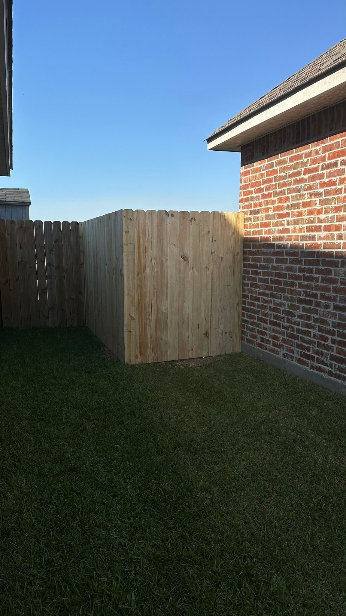 Backyard with a wooden fence, grassy lawn, and a brick house wall under clear blue sky
