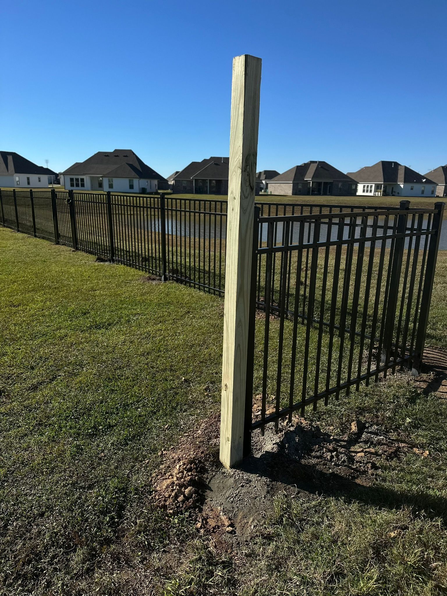 Wooden fence post beside black metal fencing in a grassy suburban yard under a clear blue sky