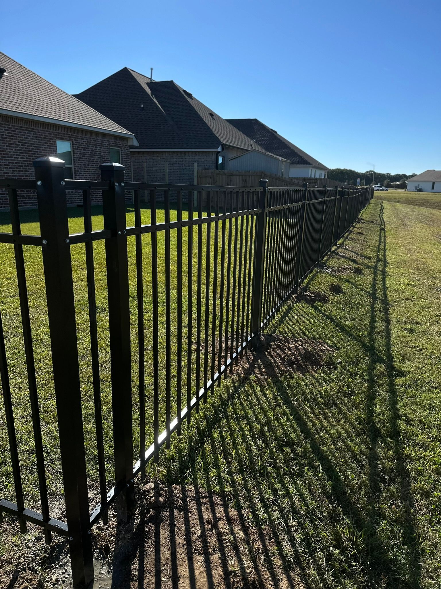 Black metal fence beside green grass and suburban houses under a clear blue sky.