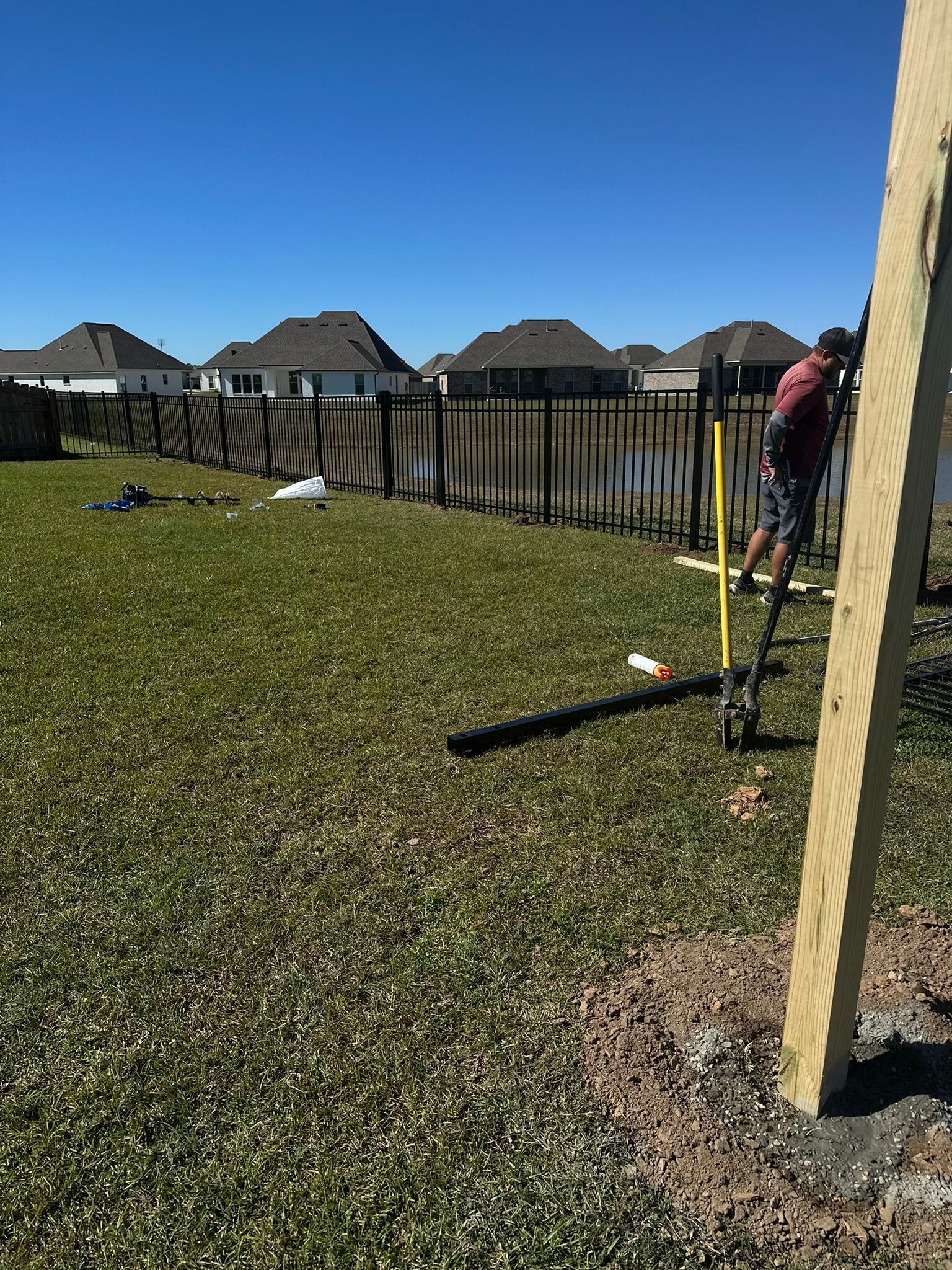 Backyard with black fence, grassy yard, and a person near a wooden post under a clear blue sky