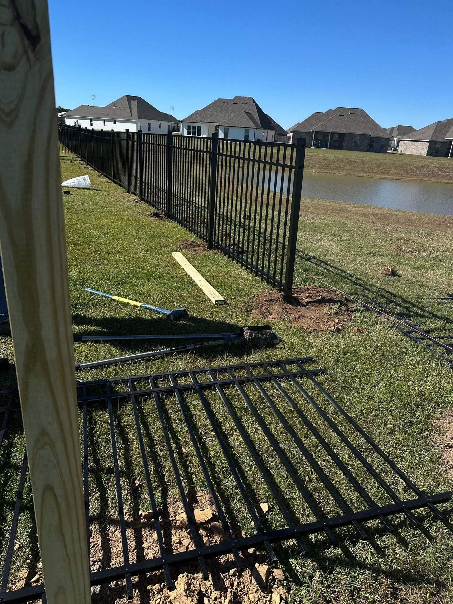 Bent black metal fence panel leaning on grass beside a pond in a sunny suburban yard