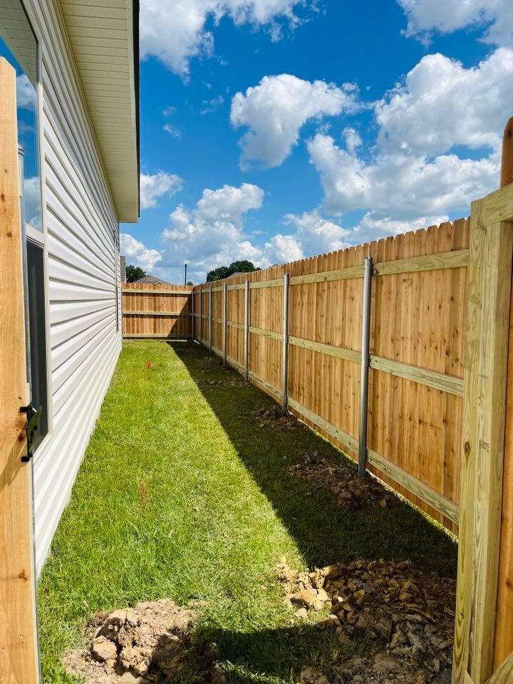 Narrow grassy side yard between a white house and wooden fence under a bright blue sky with clouds