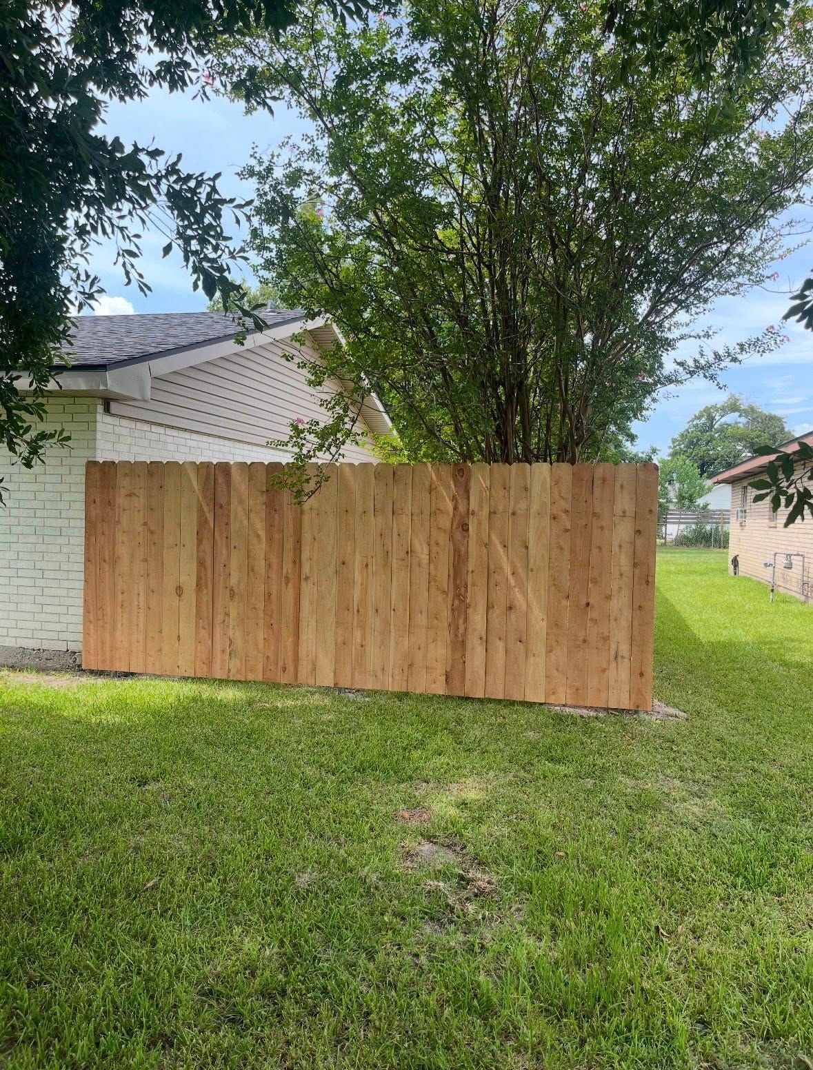 Wooden privacy fence beside a house and trees in a grassy backyard.